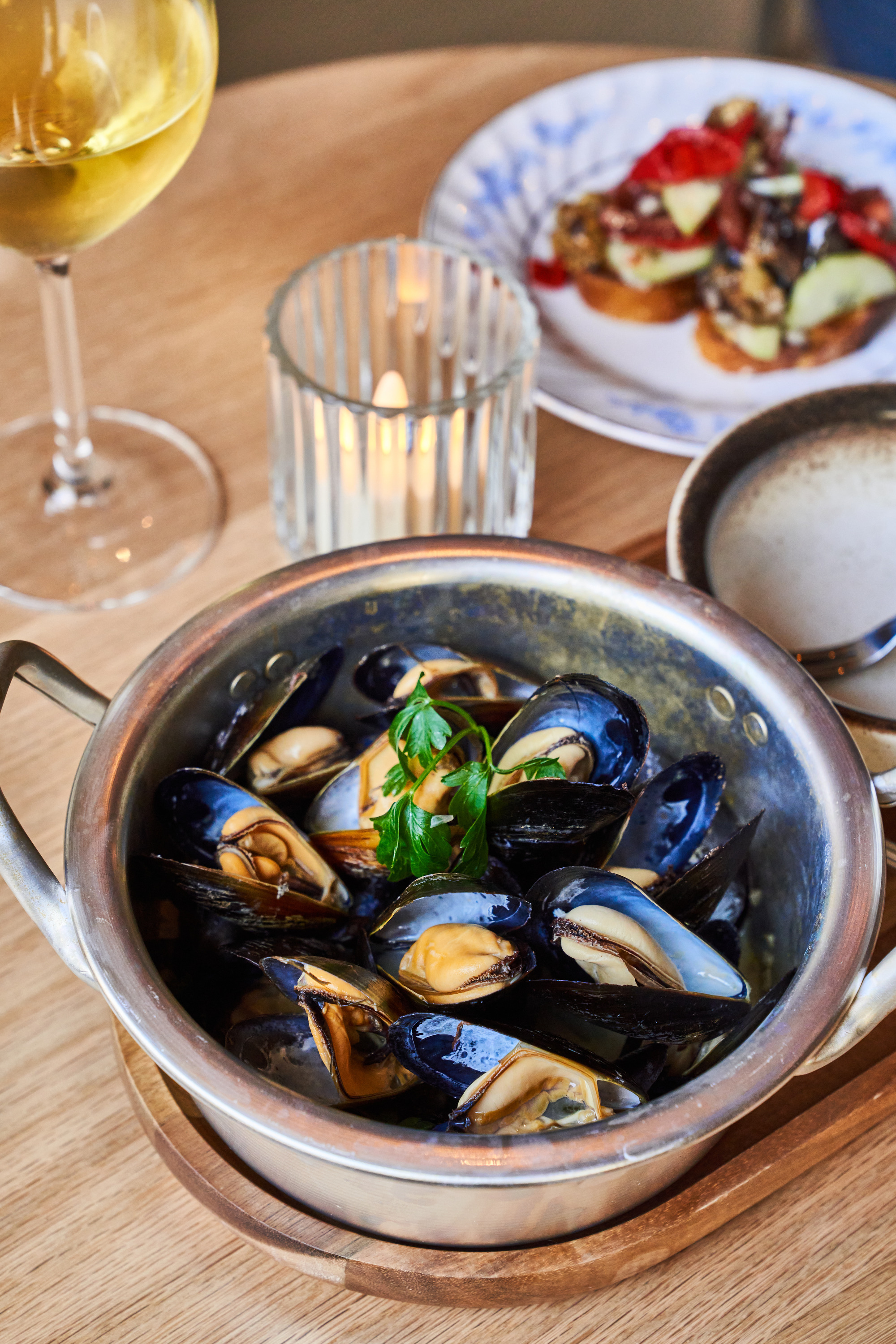 A pot of cooked mussels garnished with parsley sits on a table with a lit candle, a glass of white wine, and a plate of appetizer bruschetta.