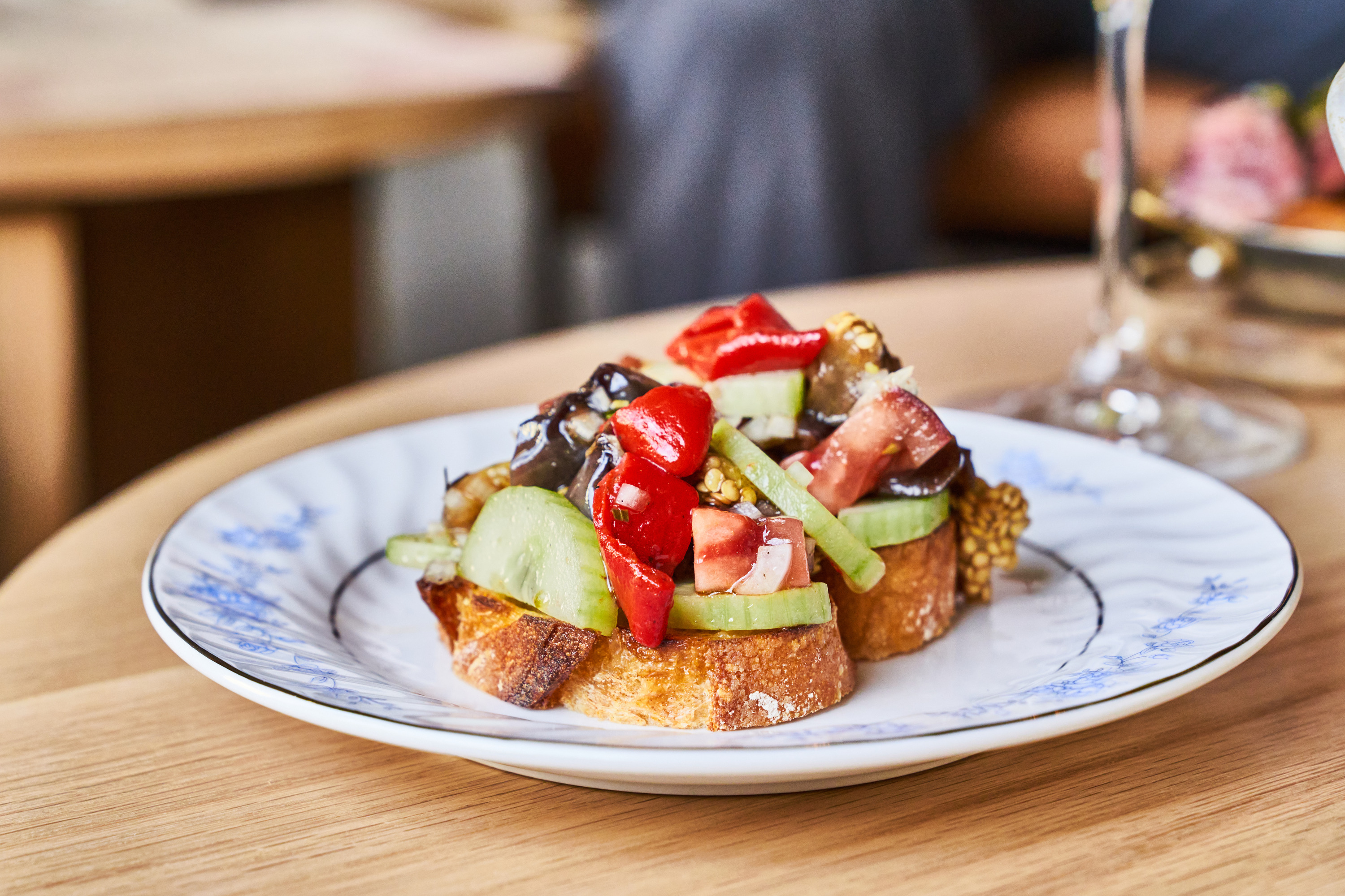 The image shows two slices of bread topped with cucumber, tomato, red peppers, and eggplant on a white plate with blue floral patterns.