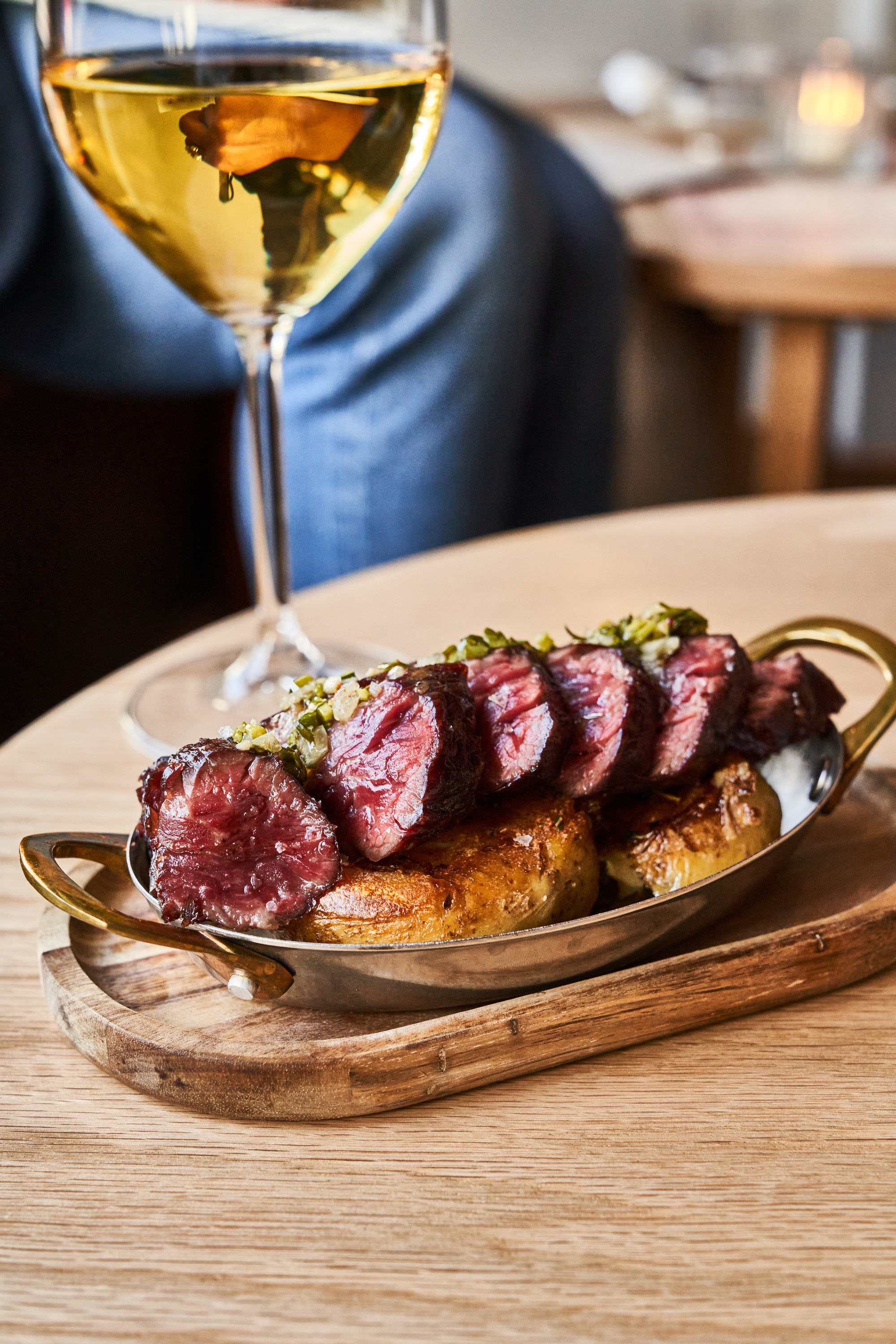 A wooden table holds a metal dish with sliced steak atop roasted potatoes. Beside it, a glass of white wine reflects a hand holding a key, with blue jeans in the background.
