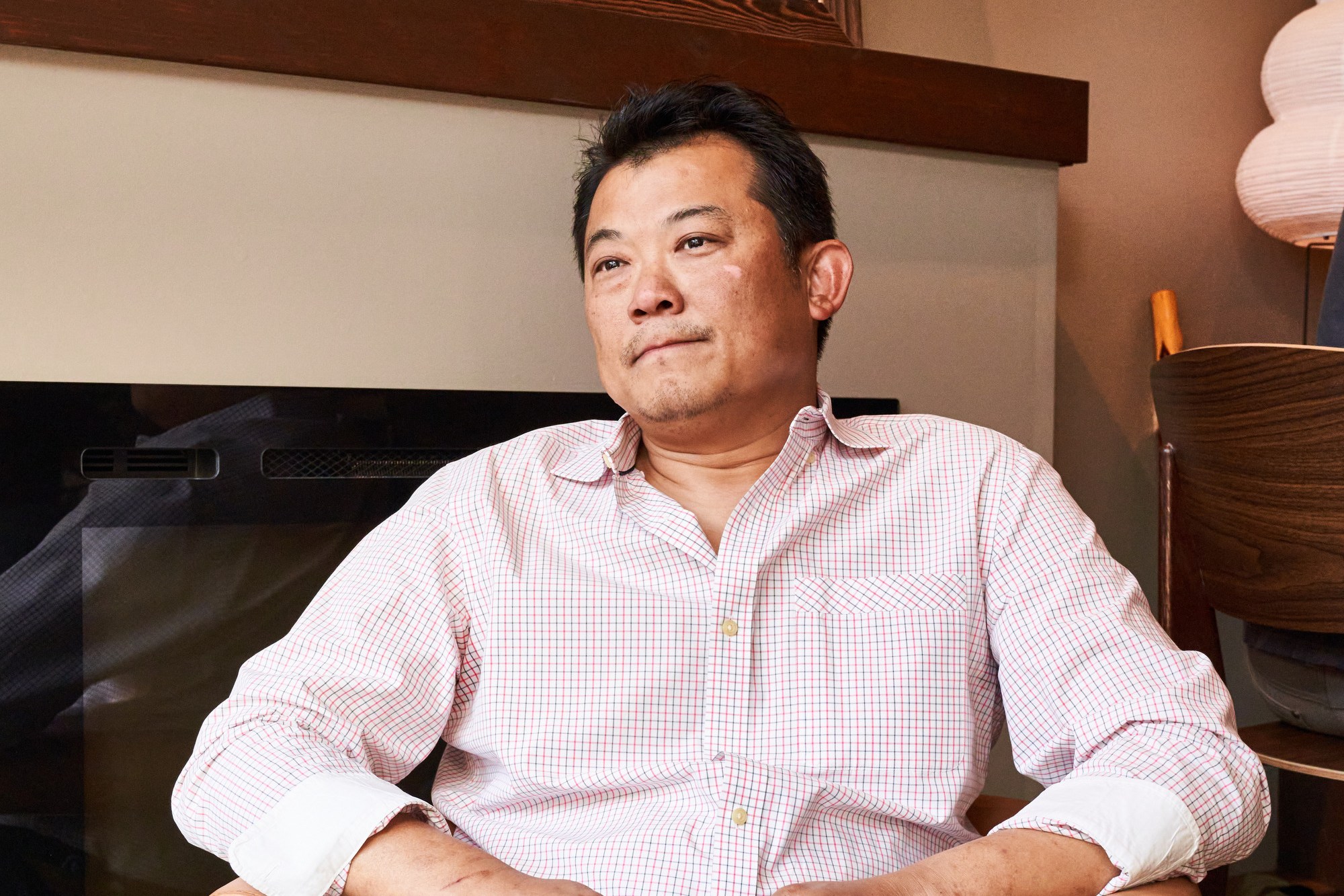 A man in a checked shirt sits in a cozy room holding a mug. Behind him, there's a chalkboard menu listing seafood items and prices.