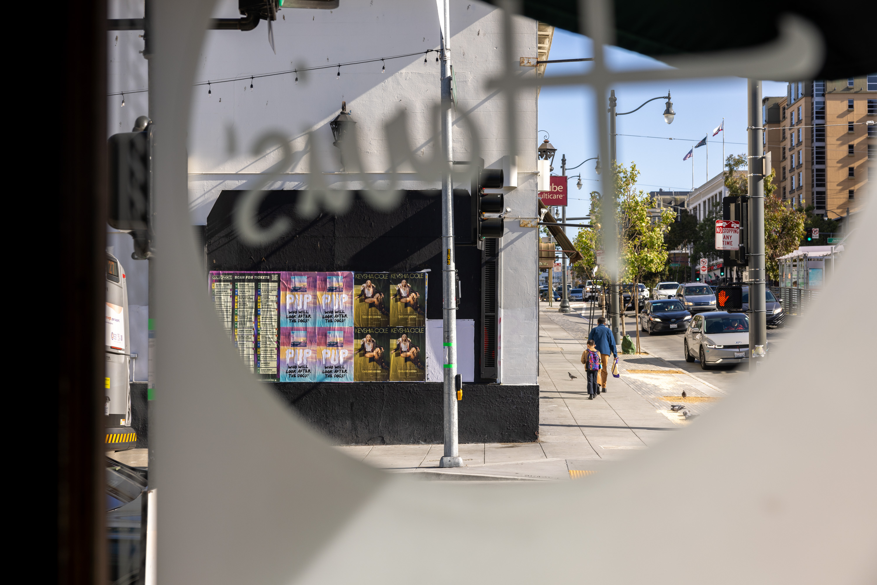 A city street view through a circular window, showing people walking, cars, and posters on a building. Traffic lights and trees are visible under a clear sky.