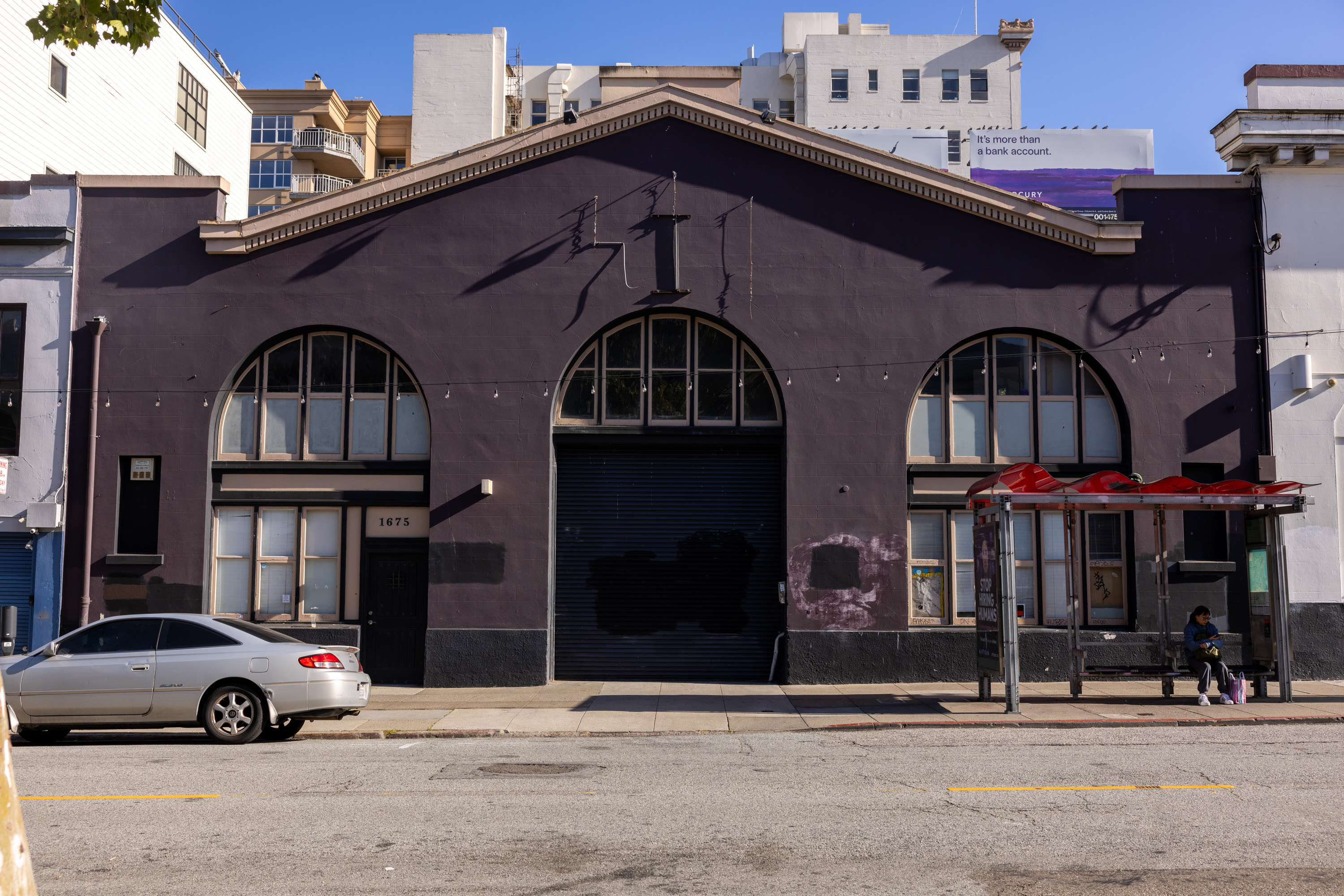A purple building with arched windows sits on a street. In front, there's a silver car and a bus stop with a person seated under a red roof.