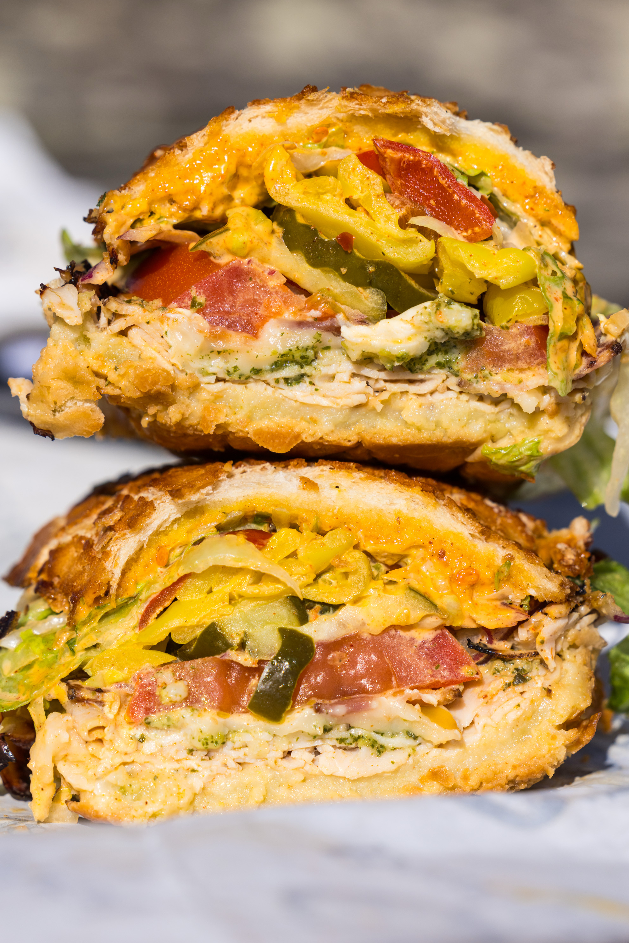 A sandwich cut in half, stacked with chicken, lettuce, tomatoes, cheddar, and pickles inside crusty bread, drizzled with a sauce.