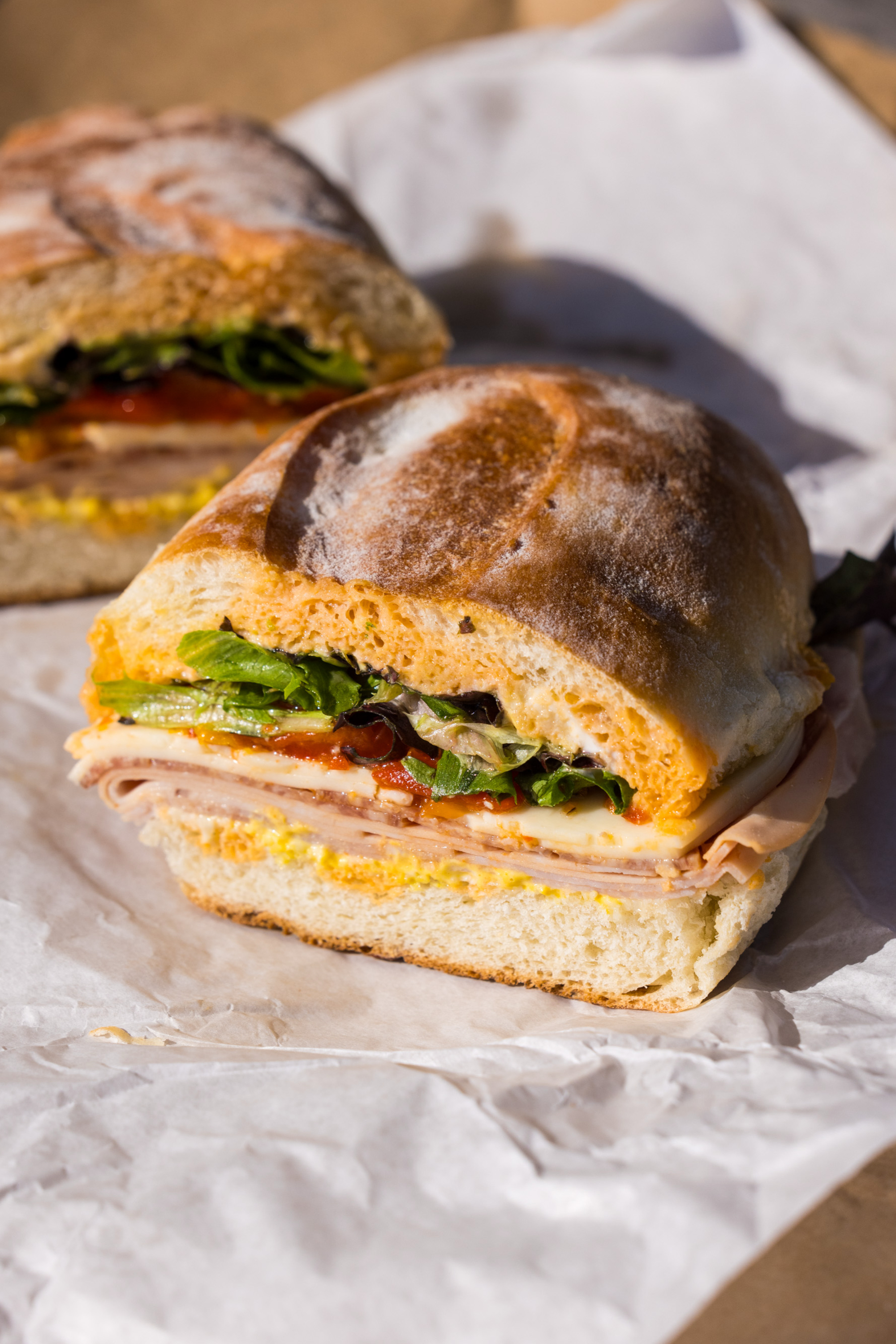 A sandwich is cut in half on white paper. It has turkey, cheese, lettuce, tomato, and mustard between two slices of crusty bread.