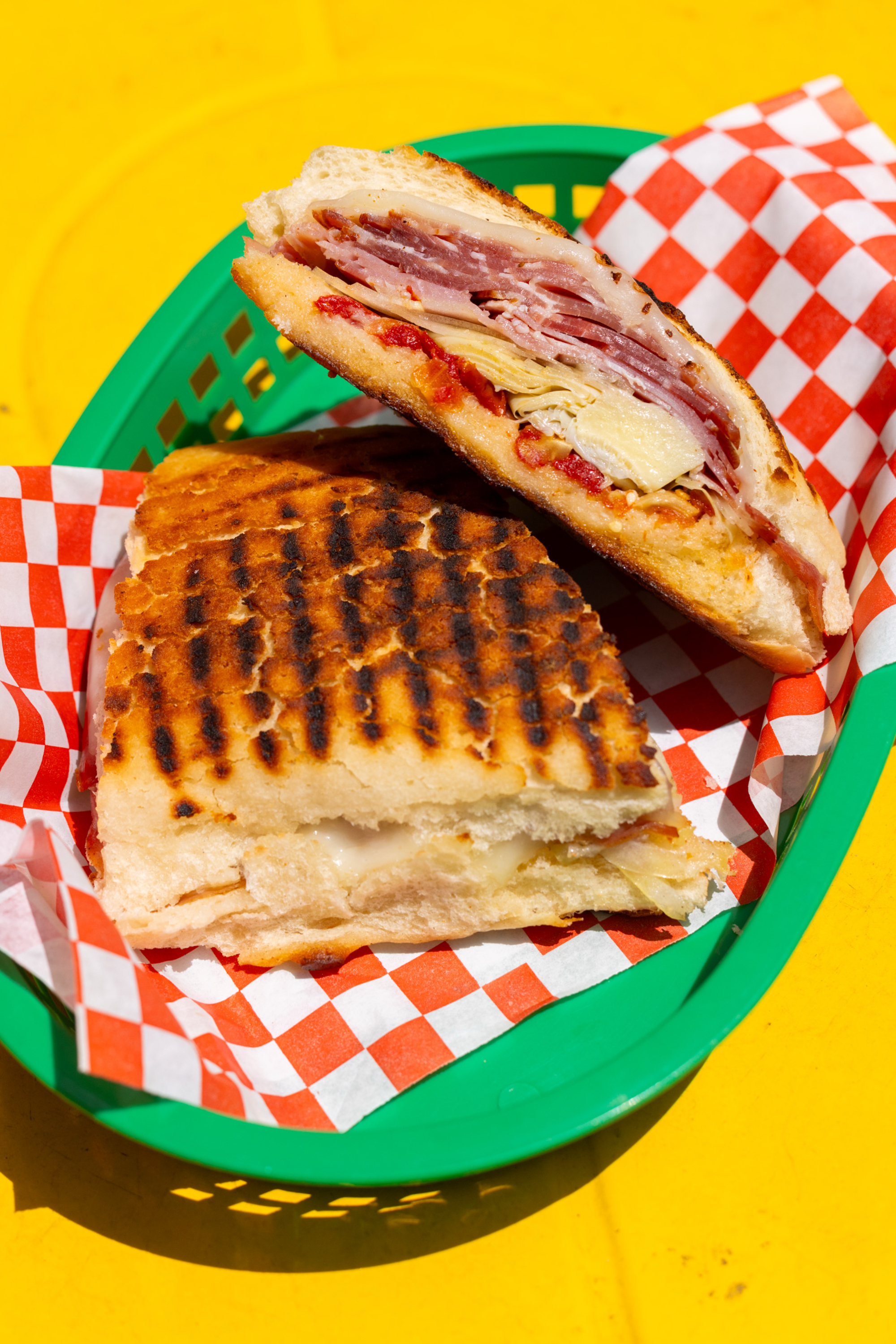 A toasted sandwich with layers of meat and cheese is placed in a green basket lined with red-and-white checkered paper on a bright yellow surface.