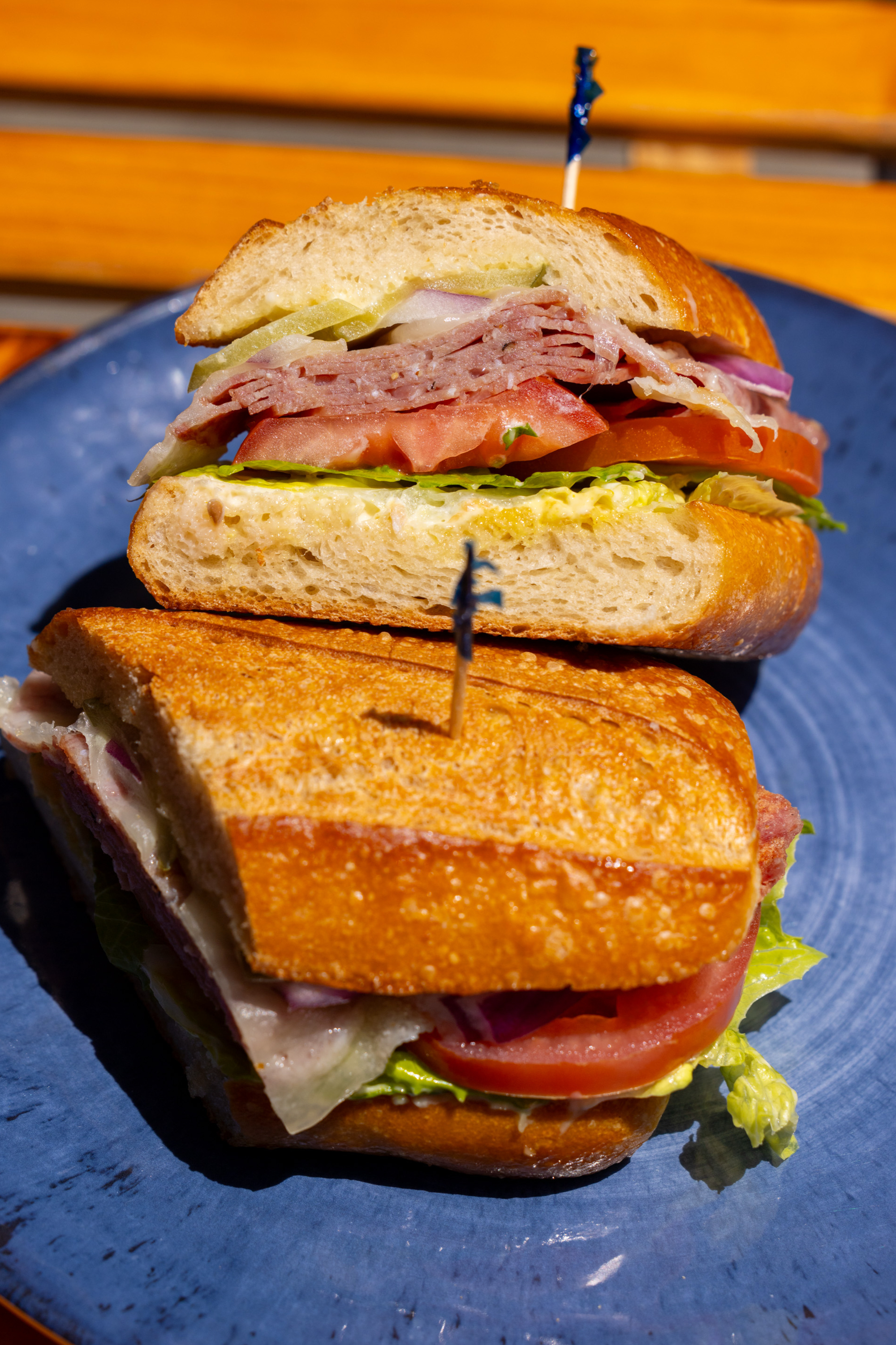 A sandwich on a blue plate with toasted bread, lettuce, tomato, sliced meats, and onions, held together with toothpicks.