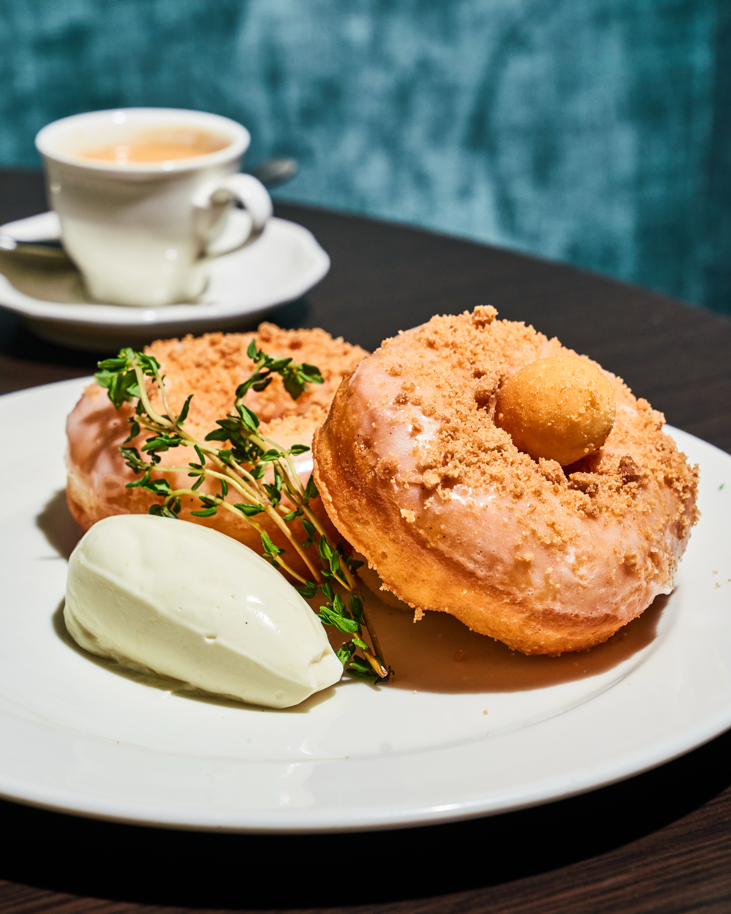 Two donuts with icing and crumbs are on a plate, accompanied by a scoop of cream and herbs. A cup of espresso sits in the background.