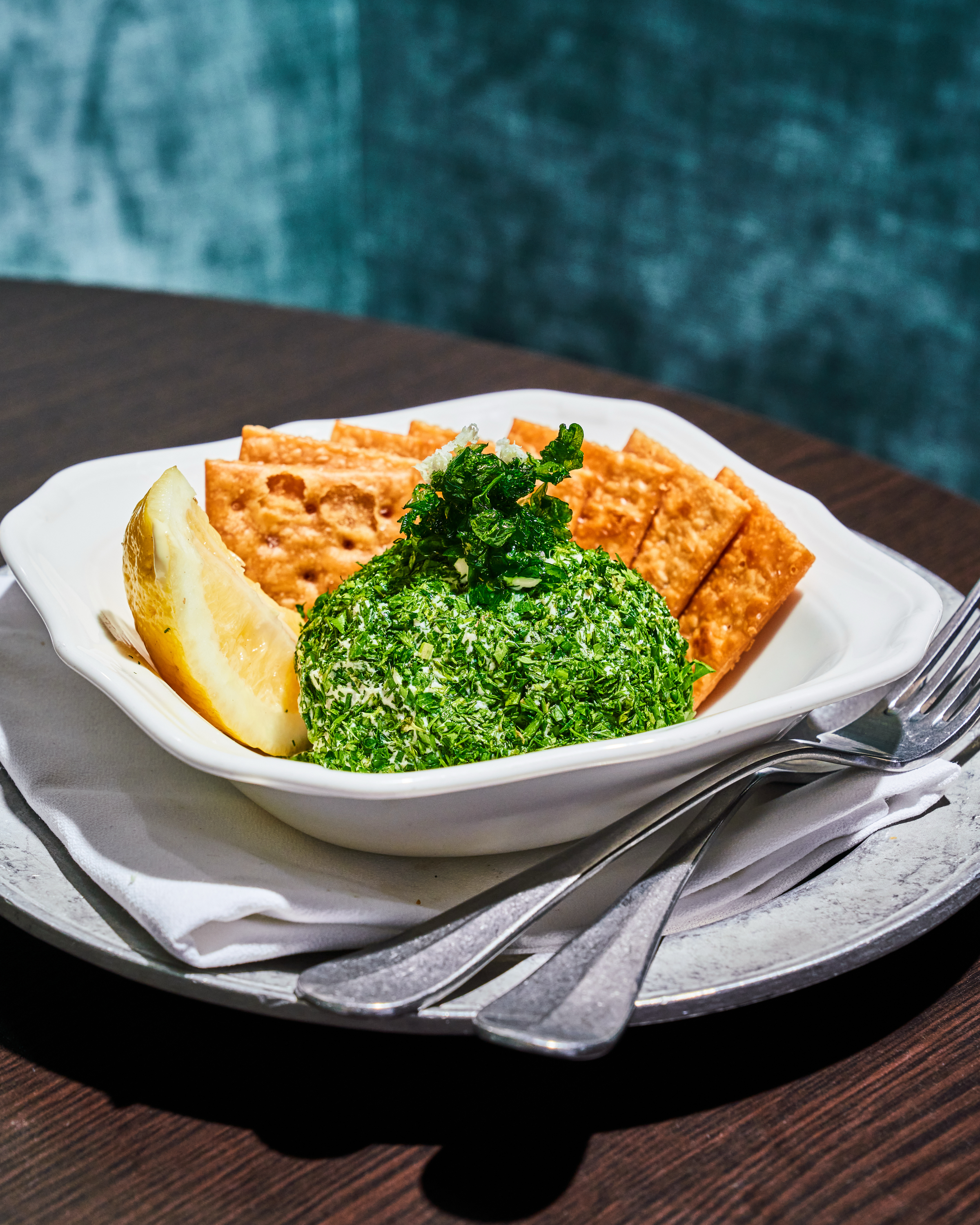 A white dish contains a green herb-covered cheese ball, a wedge of lemon, and several square crackers, set on a metal tray with a fork and knife.