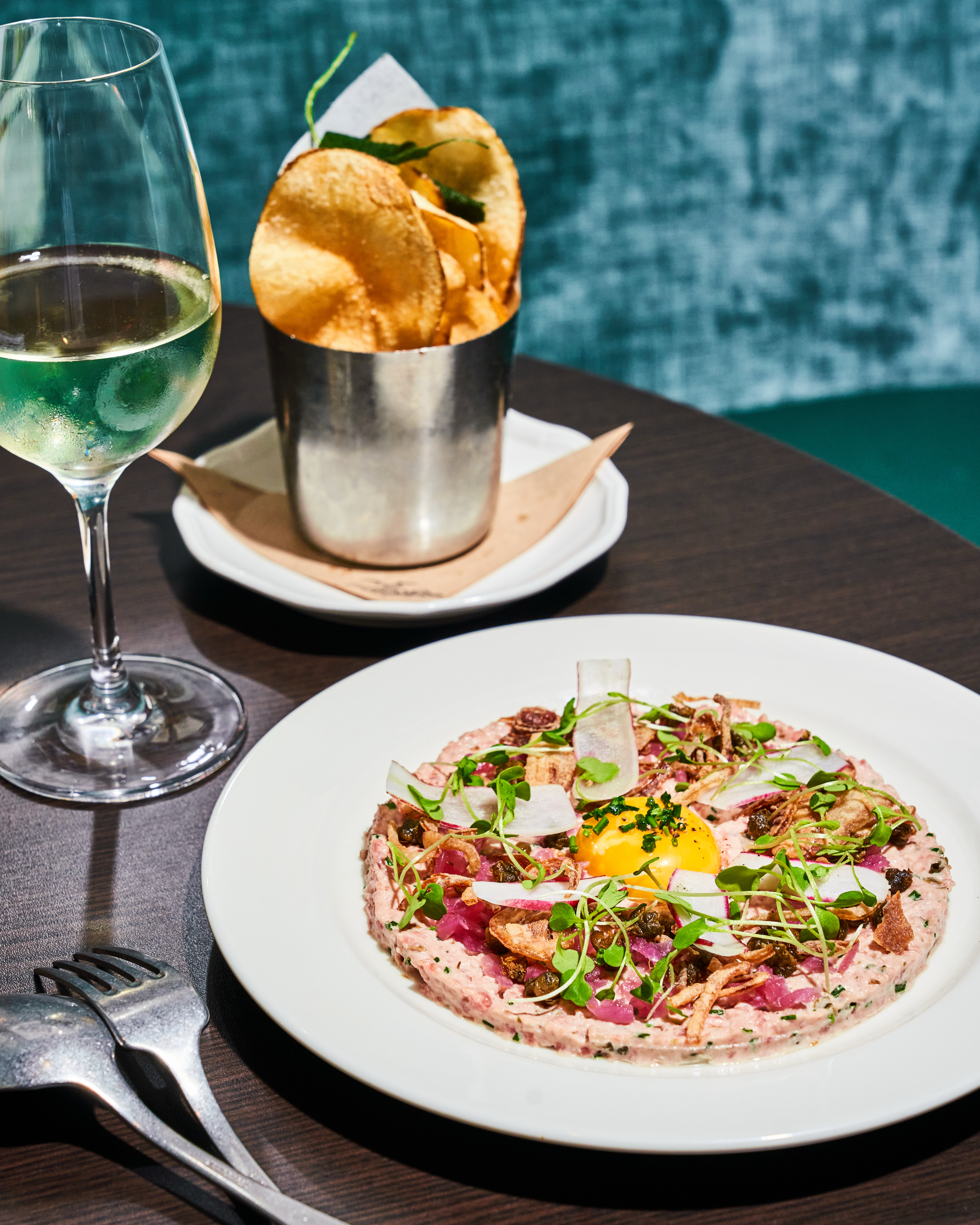 A white plate with a delicately arranged dish featuring raw beef tartare, garnished with an egg yolk, greens, and crispy elements sits next to a glass of white wine and a cup of chips.