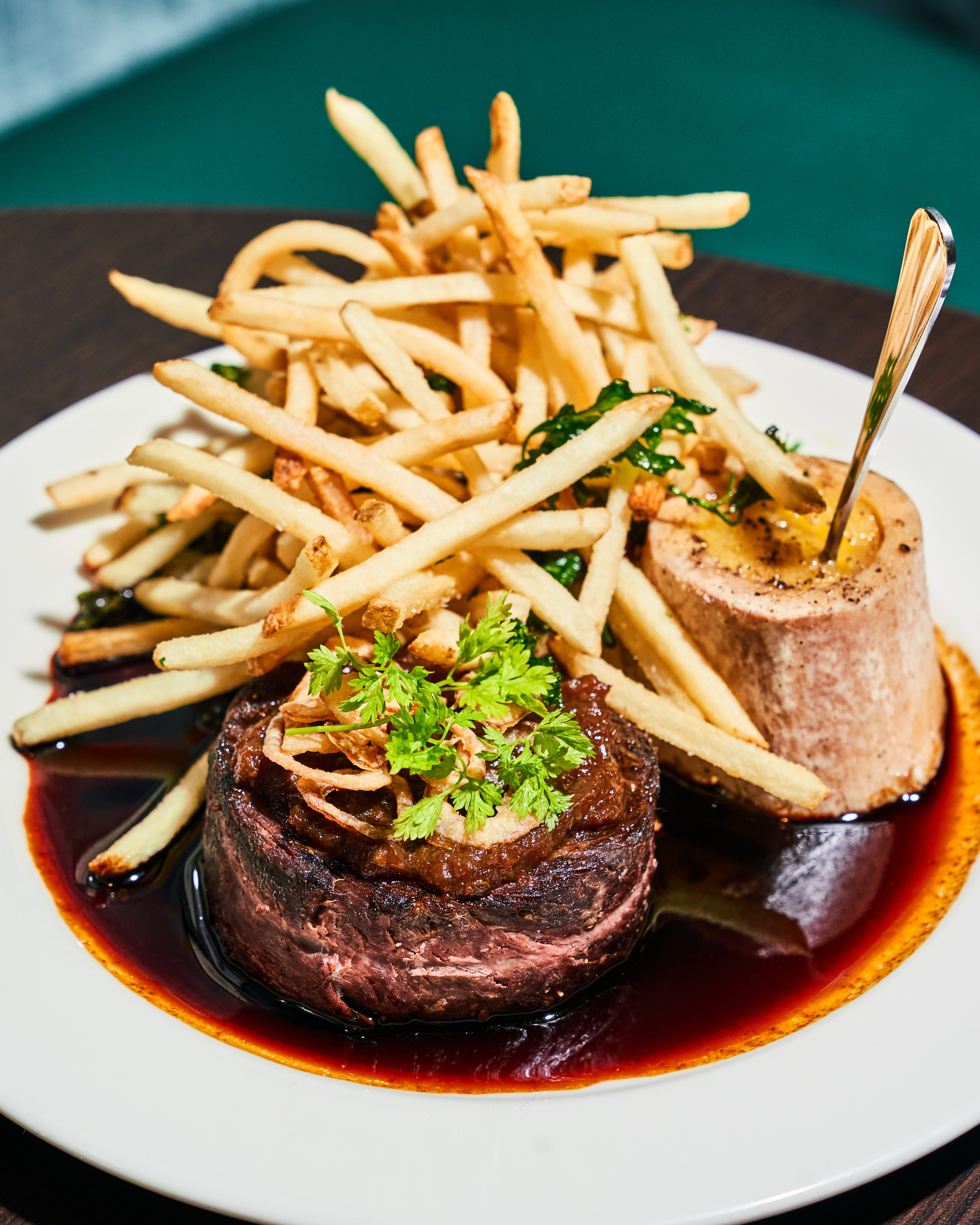 A plate features a juicy steak topped with herbs, surrounded by a rich sauce. Beside it, there's a bone marrow and a pile of crispy fries.