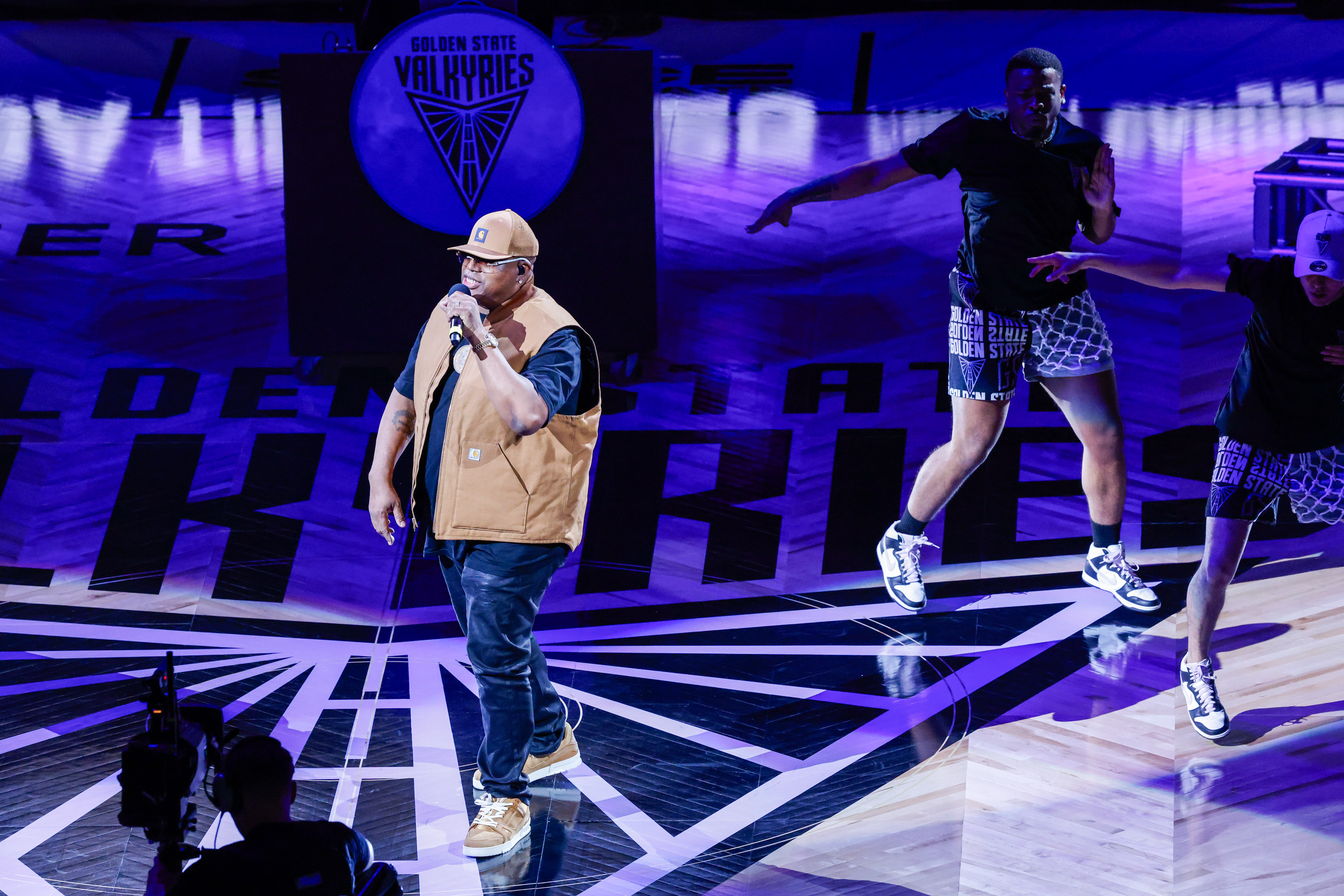 A person is performing on a stage with a microphone, wearing a tan cap and vest. Behind them, a dancer jumps, and the background has purple lighting.