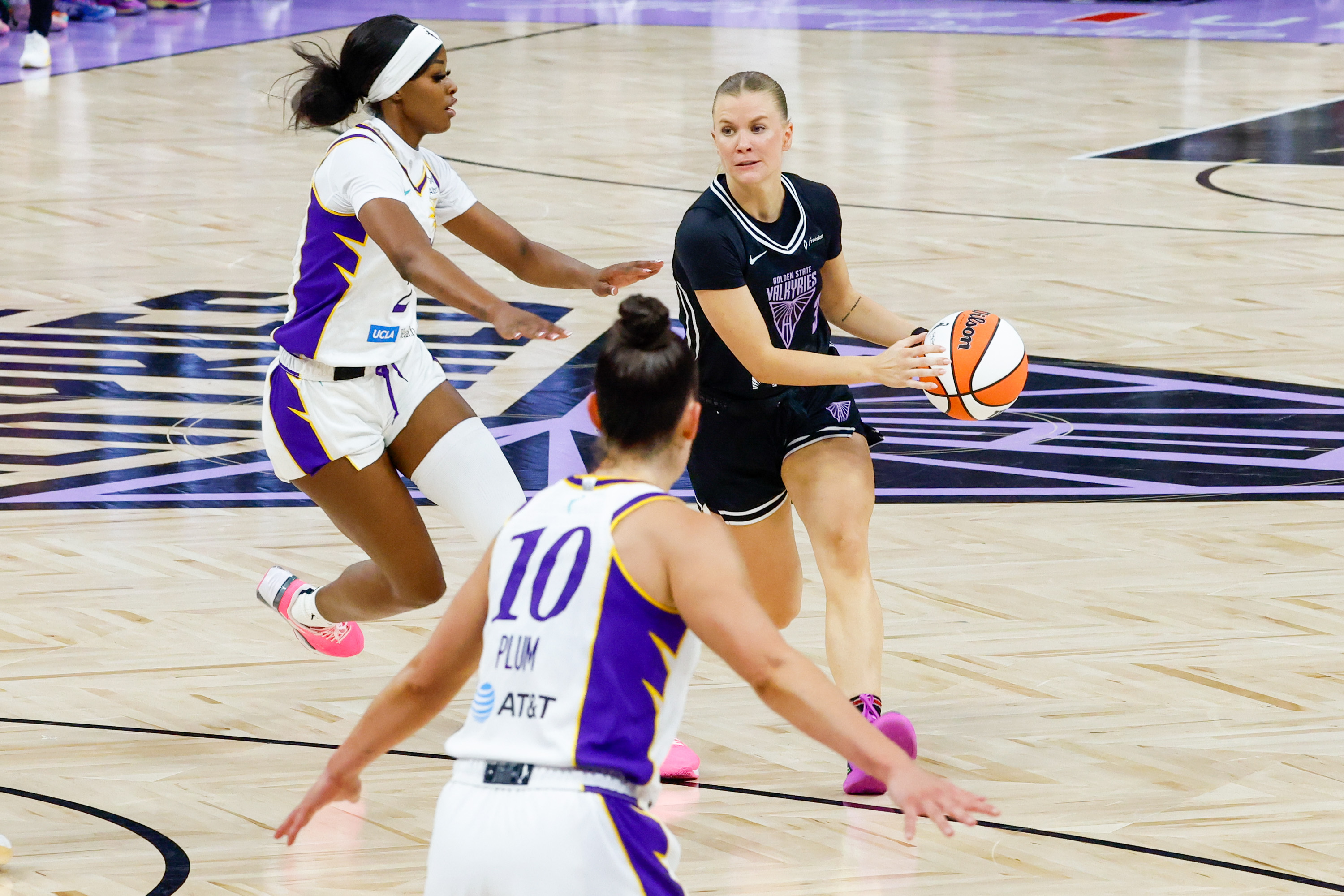 A basketball game with three players, one in black holding the ball and two in white defending. The court has light wood flooring with purple accents.