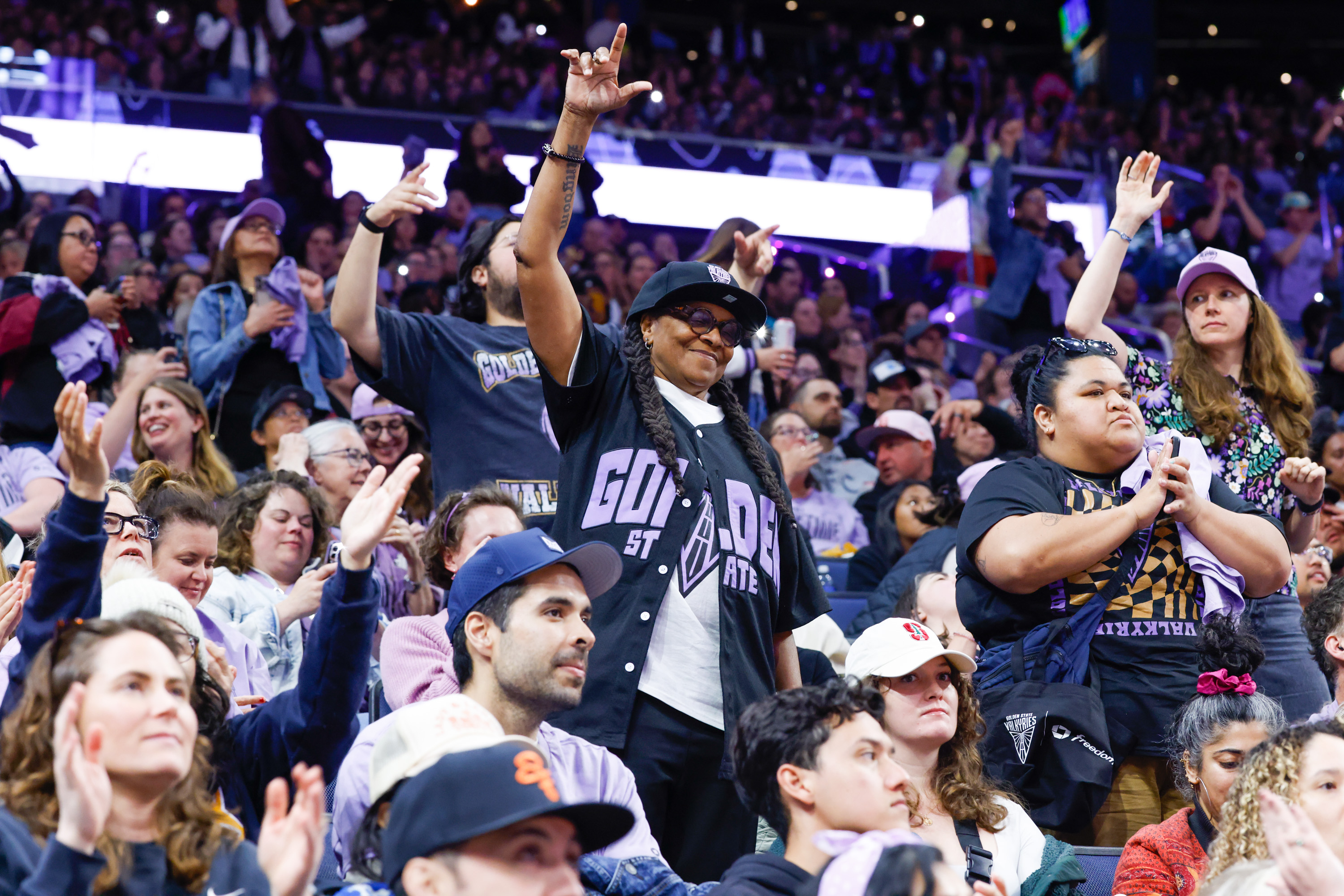 A lively crowd stands and cheers at a sports event, with fans in jerseys and hats. One person raises their hands, and others clap and take photos.
