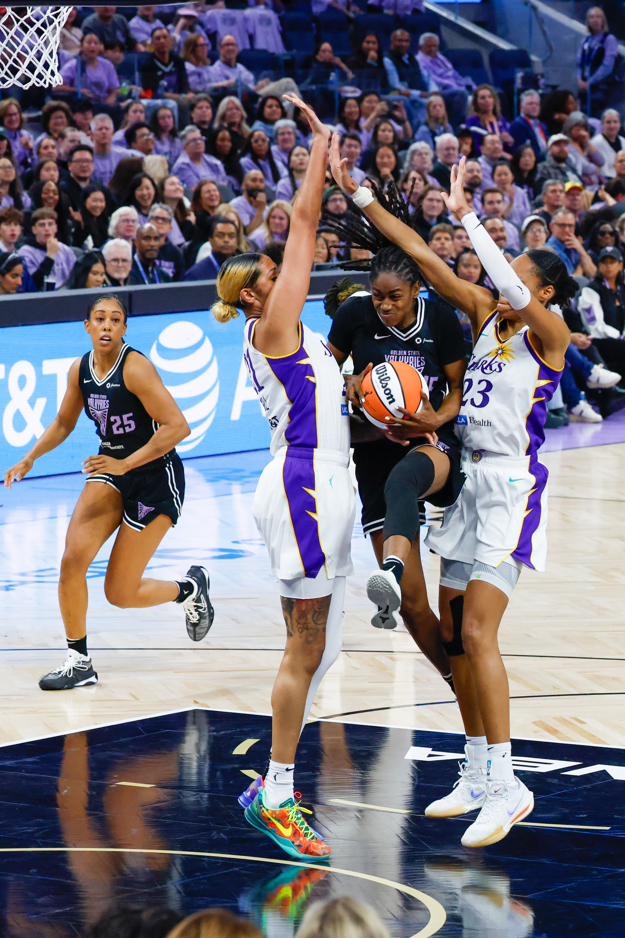 Two basketball players in white jerseys defend a player in black holding the ball, while one more player follows closely. The crowd watches intently.