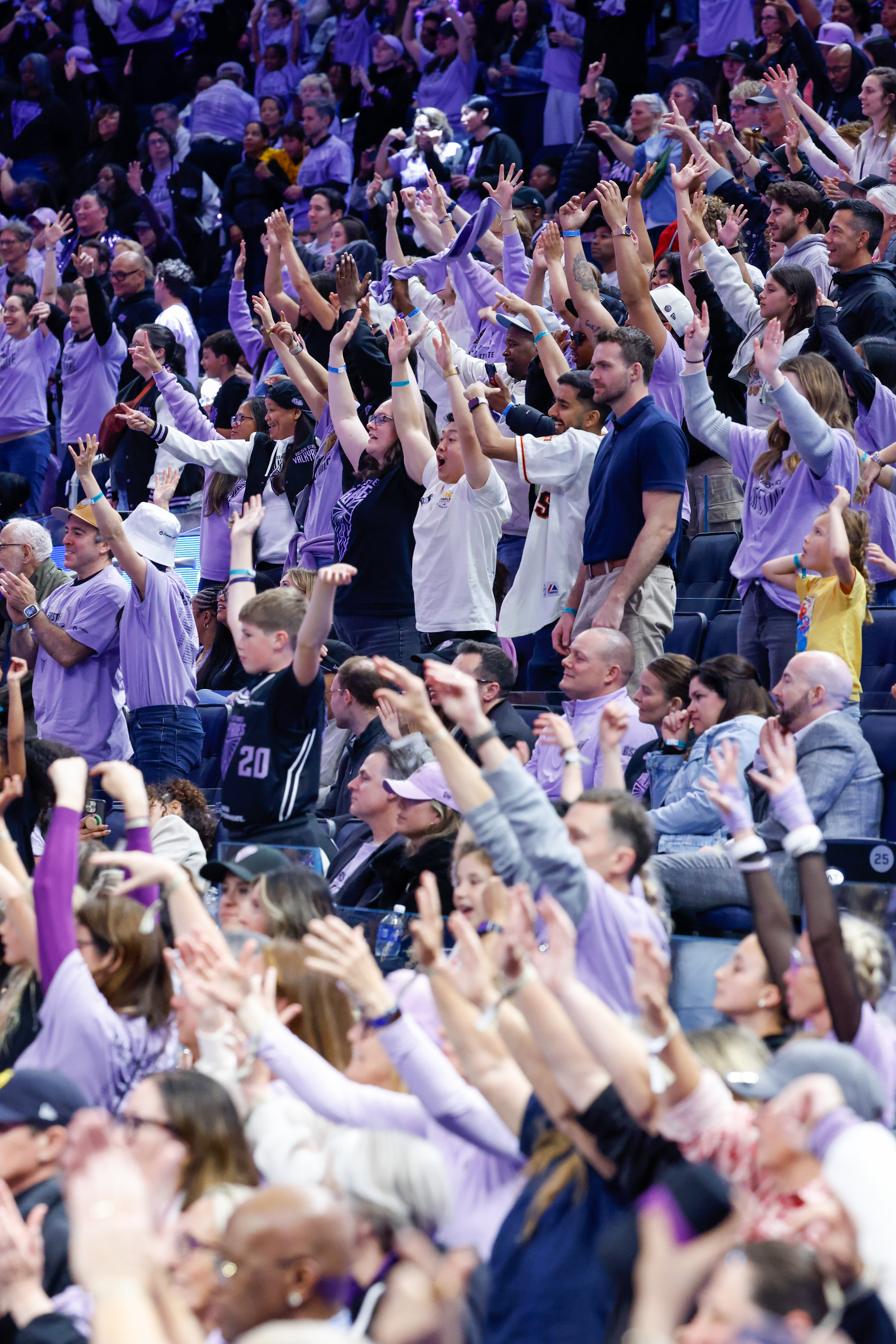 A lively crowd of people in a stadium stands with their hands raised, wearing various casual outfits, visibly excited and cheering enthusiastically.