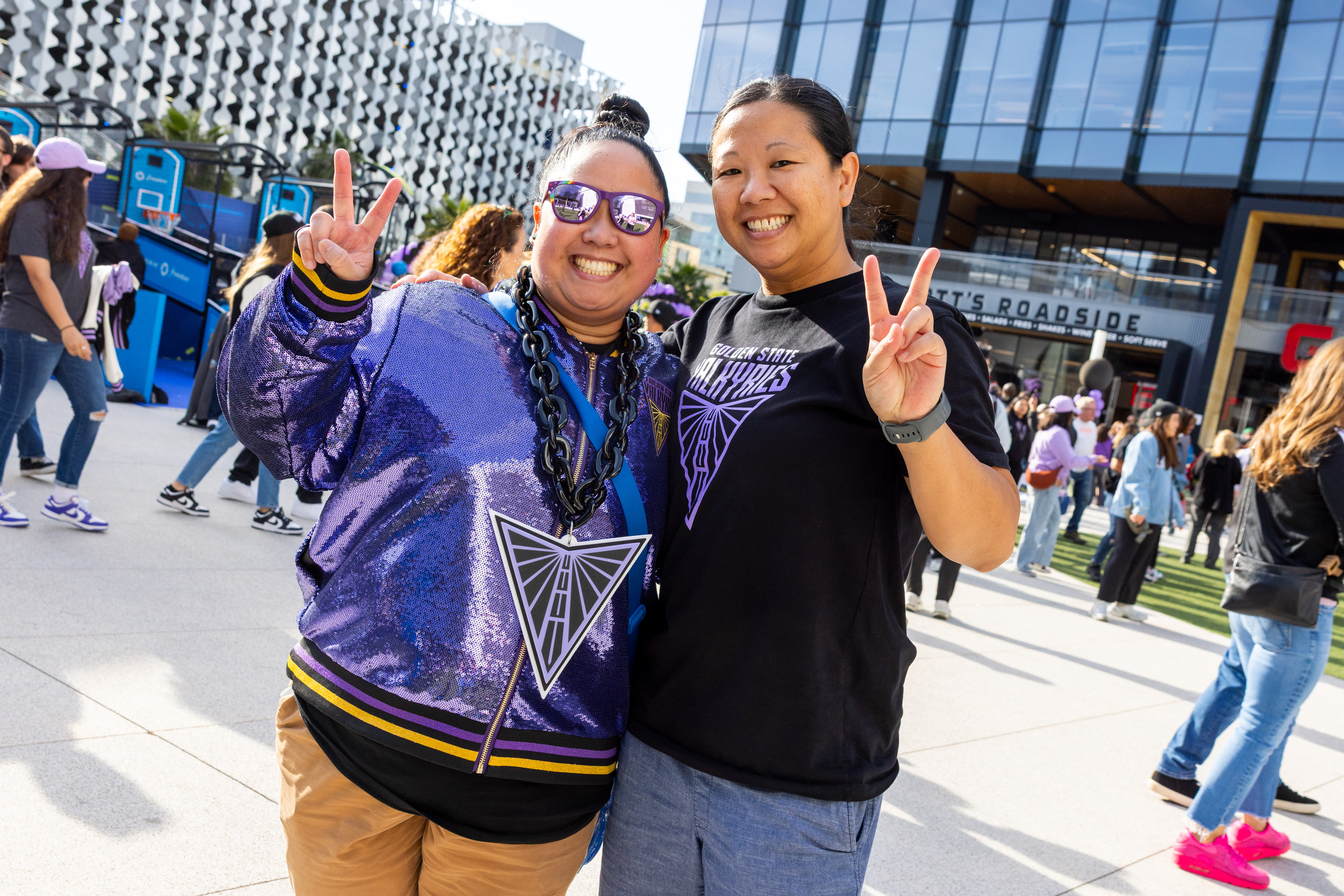 Two people pose outside, smiling and flashing peace signs. They're amidst a crowd. One wears a shiny purple jacket, the other a black shirt with a geometric logo.