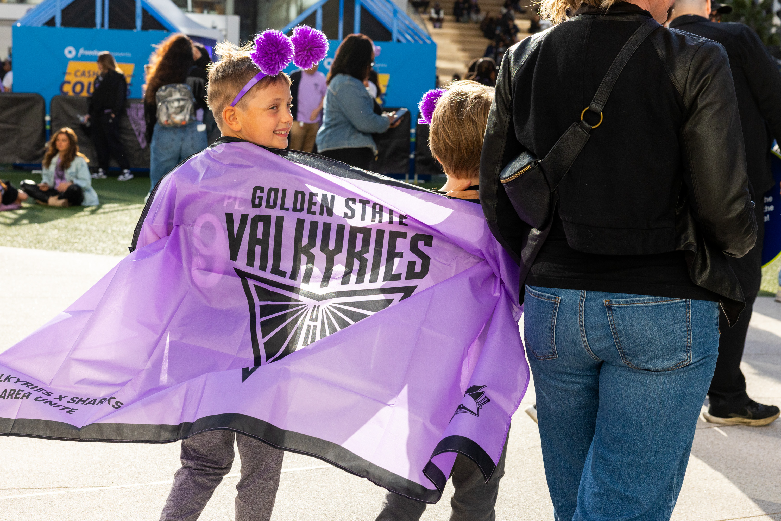 A child wearing purple headbands smiles while draped in a &quot;Golden State Valkyries&quot; banner. They're accompanied by an adult in a black jacket and jeans.