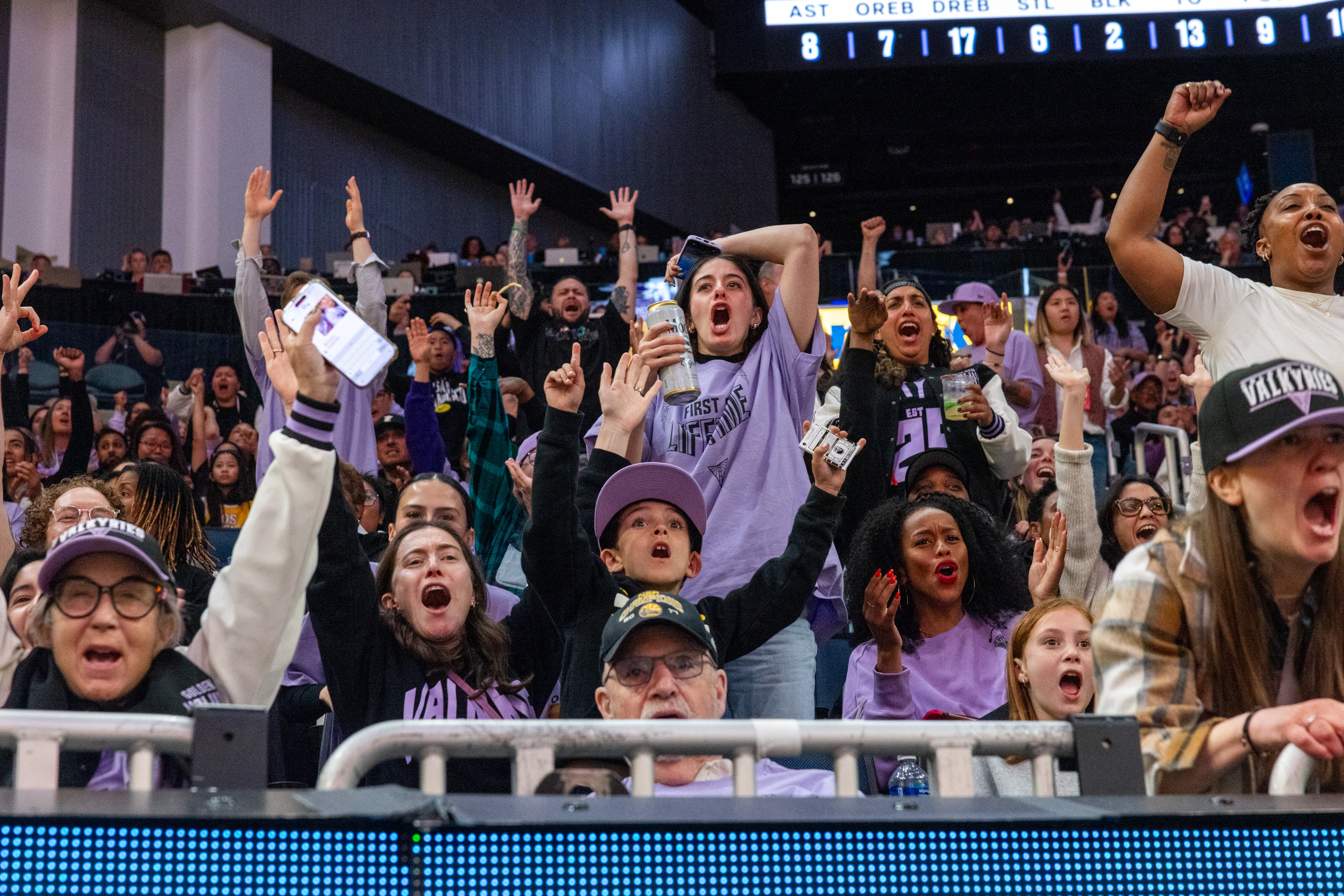 A crowd of excited sports fans is cheering wildly, with raised arms and enthusiastic expressions. Some hold drinks and phones, showing their support and energy.