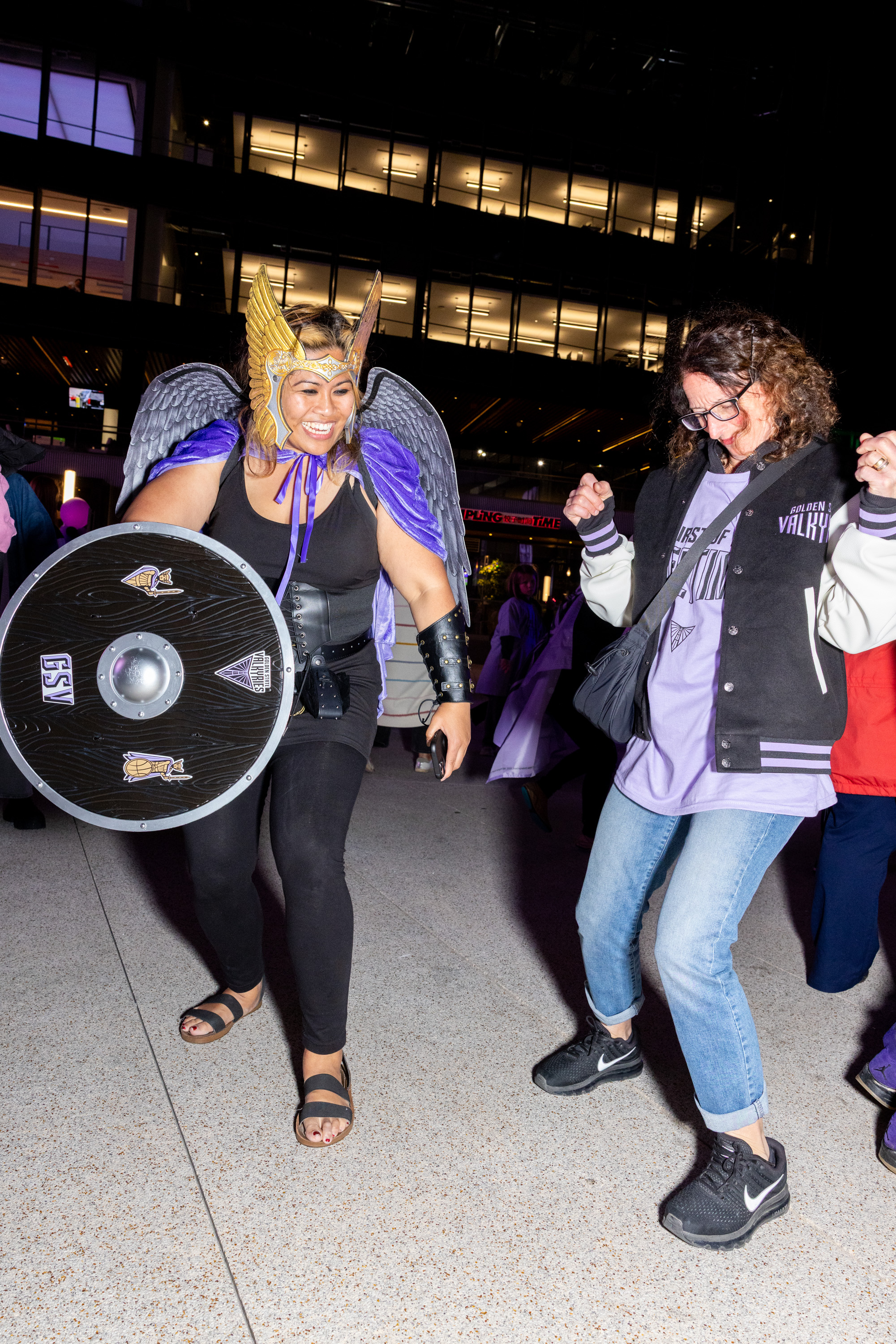 Two people are joyfully dancing outside at night. One, dressed as a warrior with a helmet and shield, wears wings and a purple cape. The other is in casual attire.