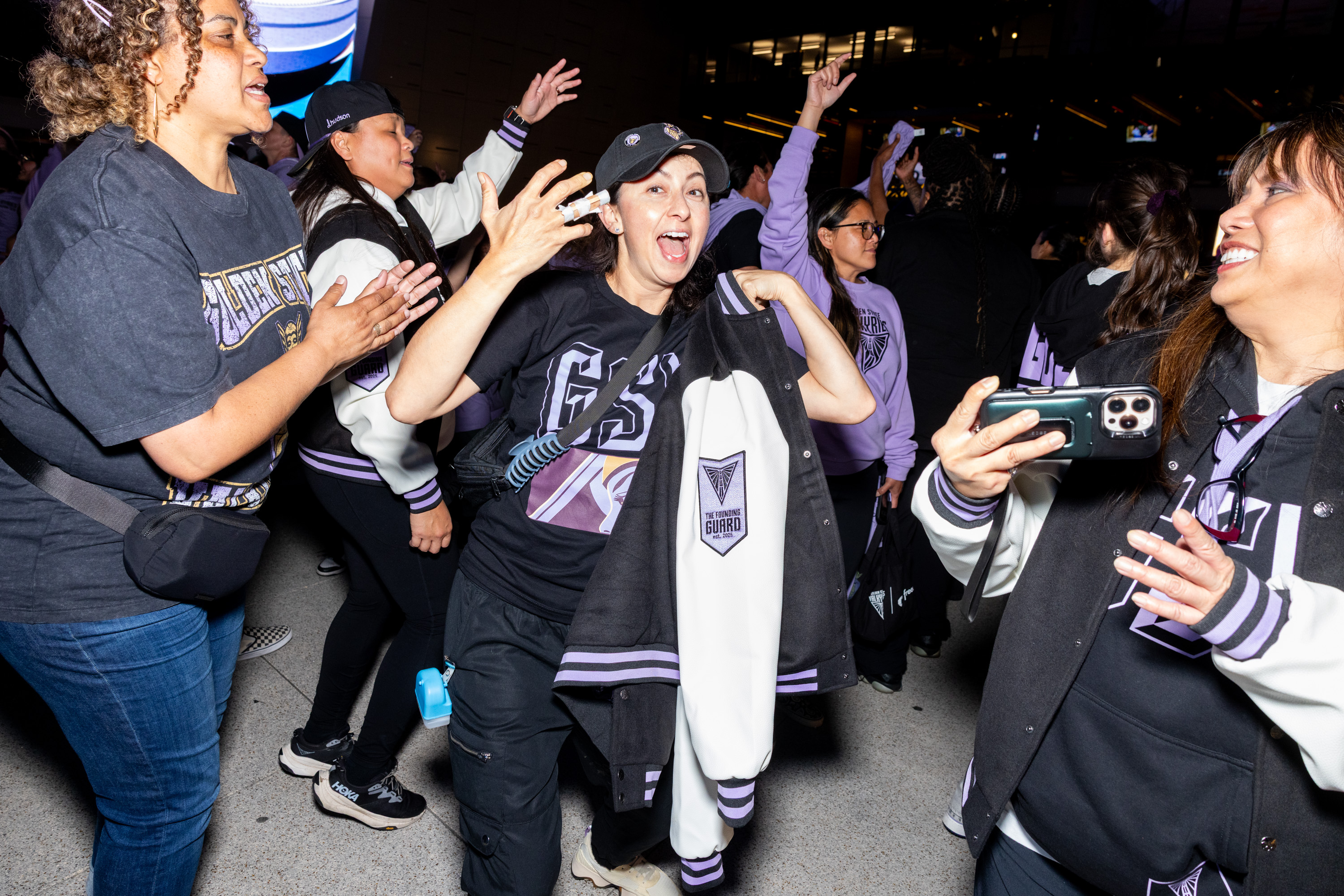 A group of enthusiastic people, mostly women, are wearing matching jackets and shirts while celebrating. One holds a jacket, and another takes a photo.