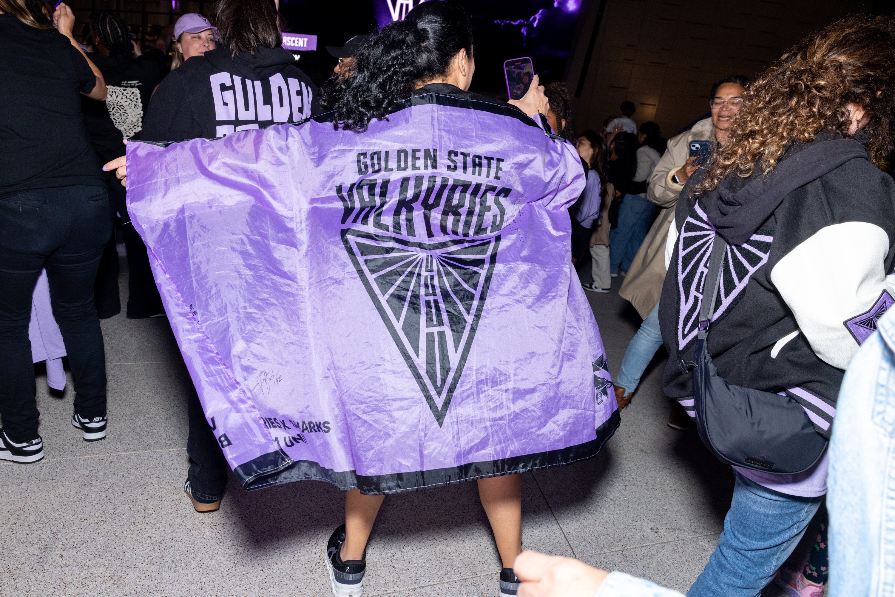 A person, seen from behind, wears a large purple jacket with "Golden State Valkyries" printed on it. They're in a crowd with some holding phones.