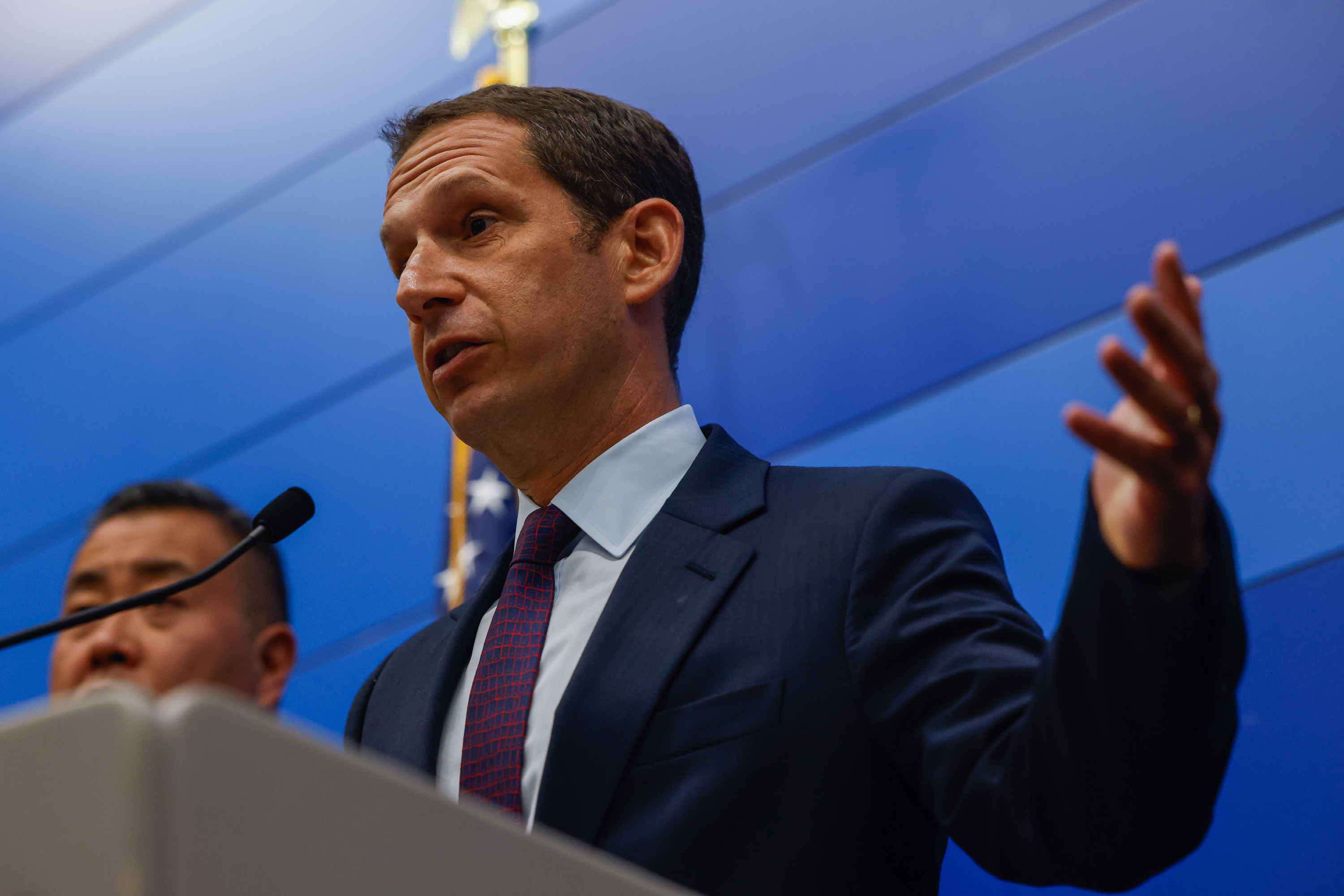 A man in a suit speaks at a podium with a microphone. Another person stands partially visible beside him. A blue wall and part of a flag are in the background.