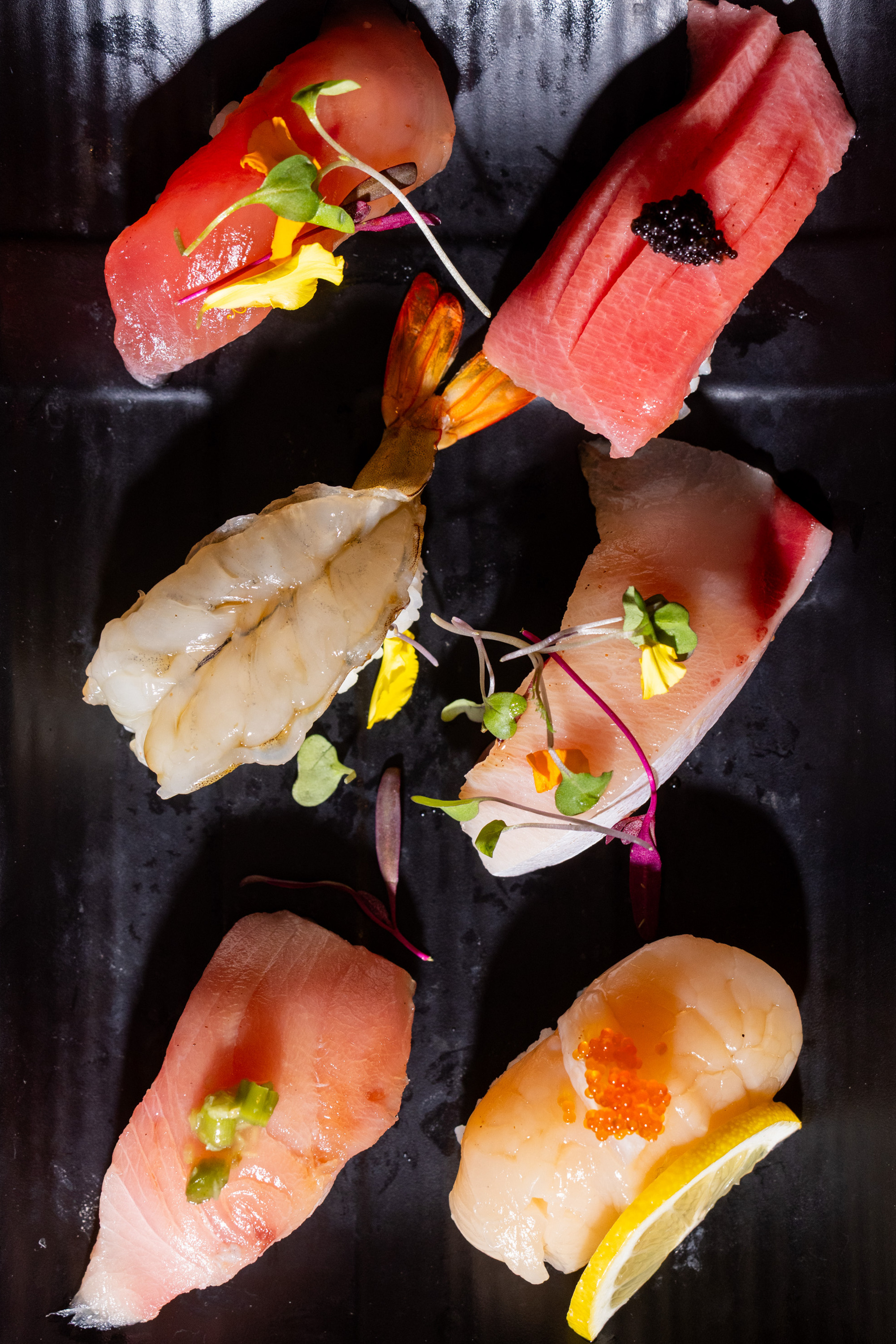 The image shows a variety of beautifully arranged sushi on a black plate, with garnishes like microgreens and a lemon slice, highlighting the fresh seafood toppings.