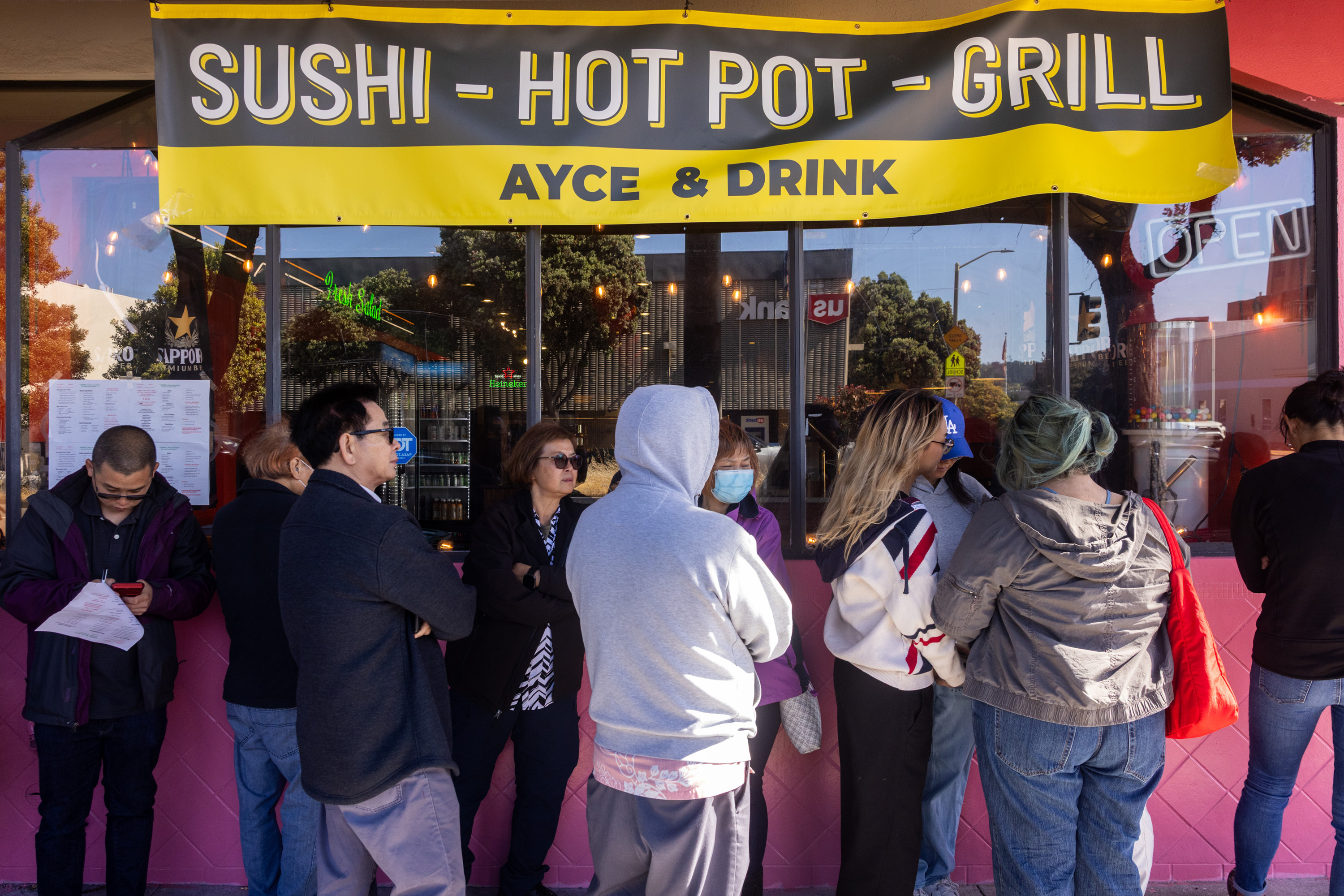People stand in line outside a restaurant with a bright yellow sign reading &quot;Sushi - Hot Pot - Grill, AYCE &amp; Drink.&quot; The scene is lively and social.