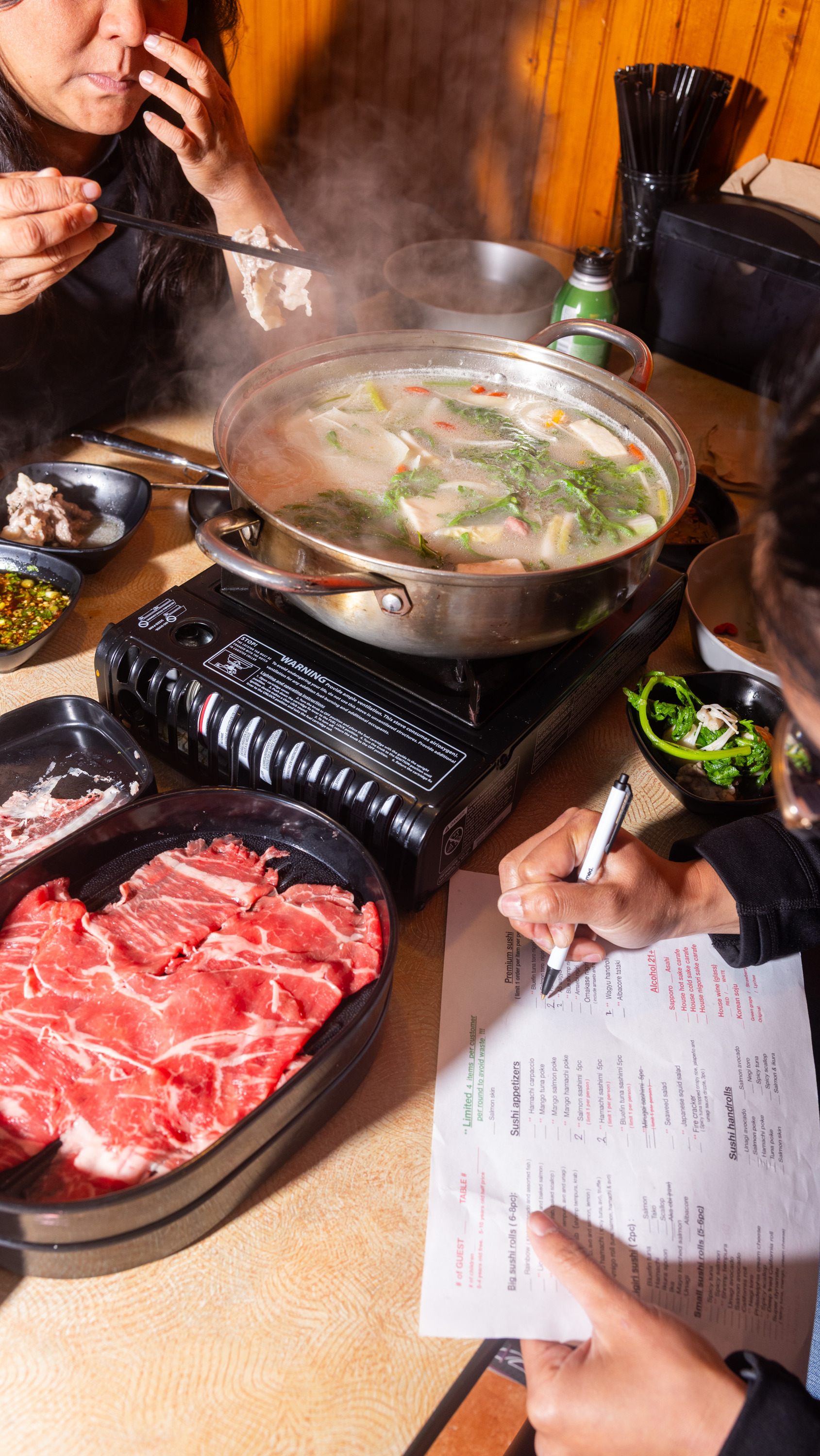 A person eats from a steaming hot pot on a table with raw meat and greens, writing on a menu. Another person holds chopsticks nearby.