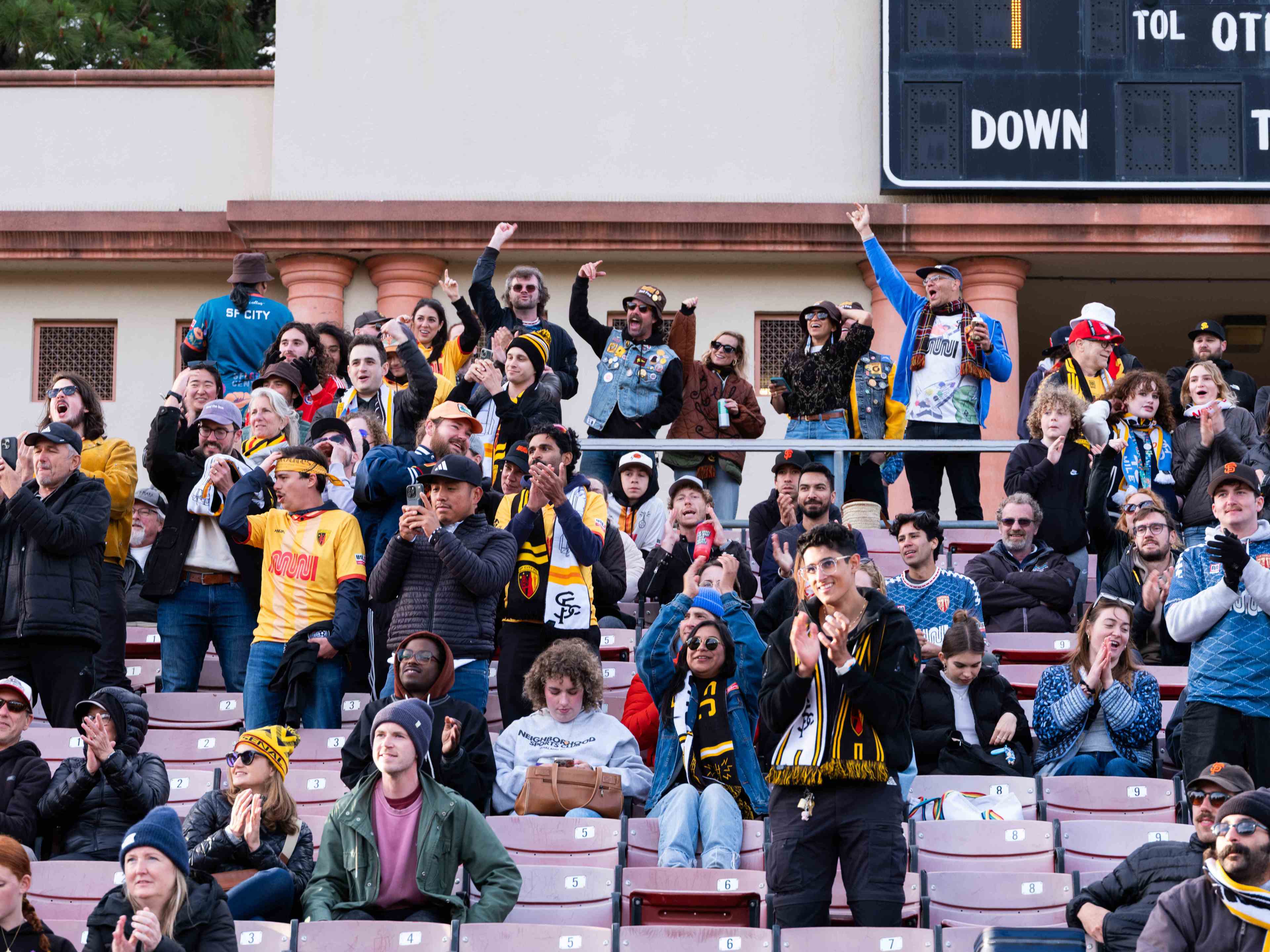 The image shows a lively crowd in a stadium, many wearing sports jerseys and scarves, cheering and clapping enthusiastically. The scoreboard is visible above them.