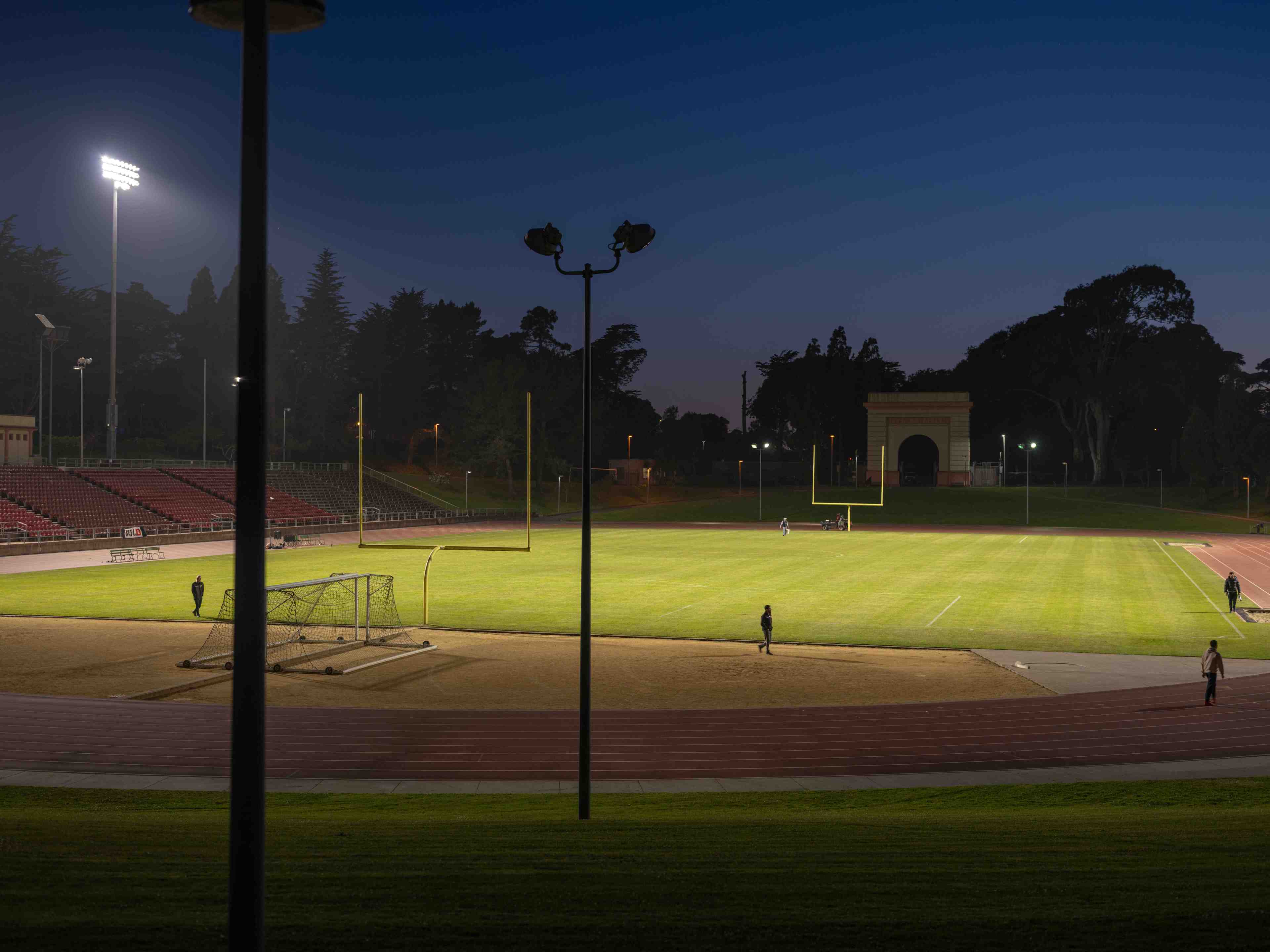 A dimly lit sports field at dusk features a running track, goalposts, and a few people on the grass. Trees and a building arch are visible in the background.