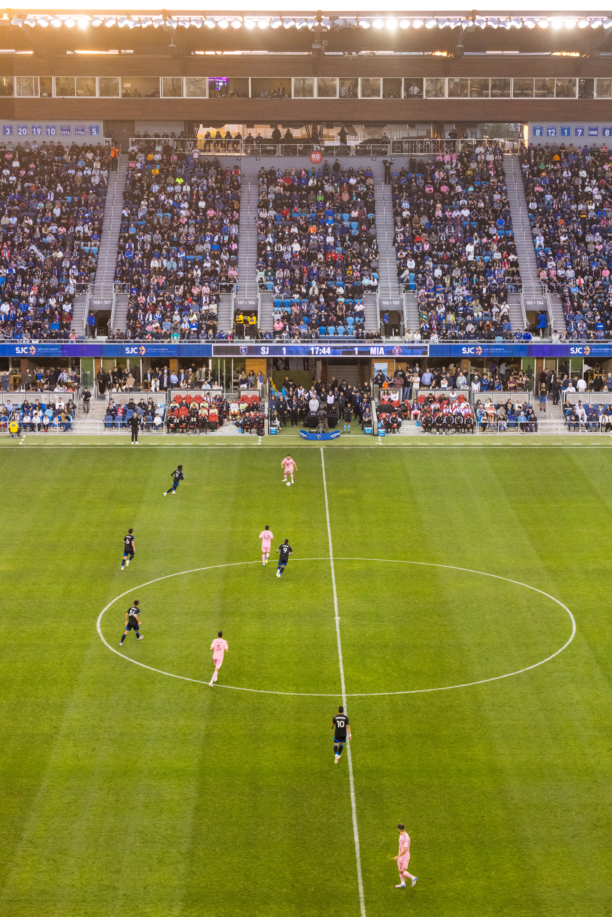 A soccer match is underway in a stadium filled with spectators. Players in pink and dark blue are on a green field, with a score of 1-1 shown on the display.