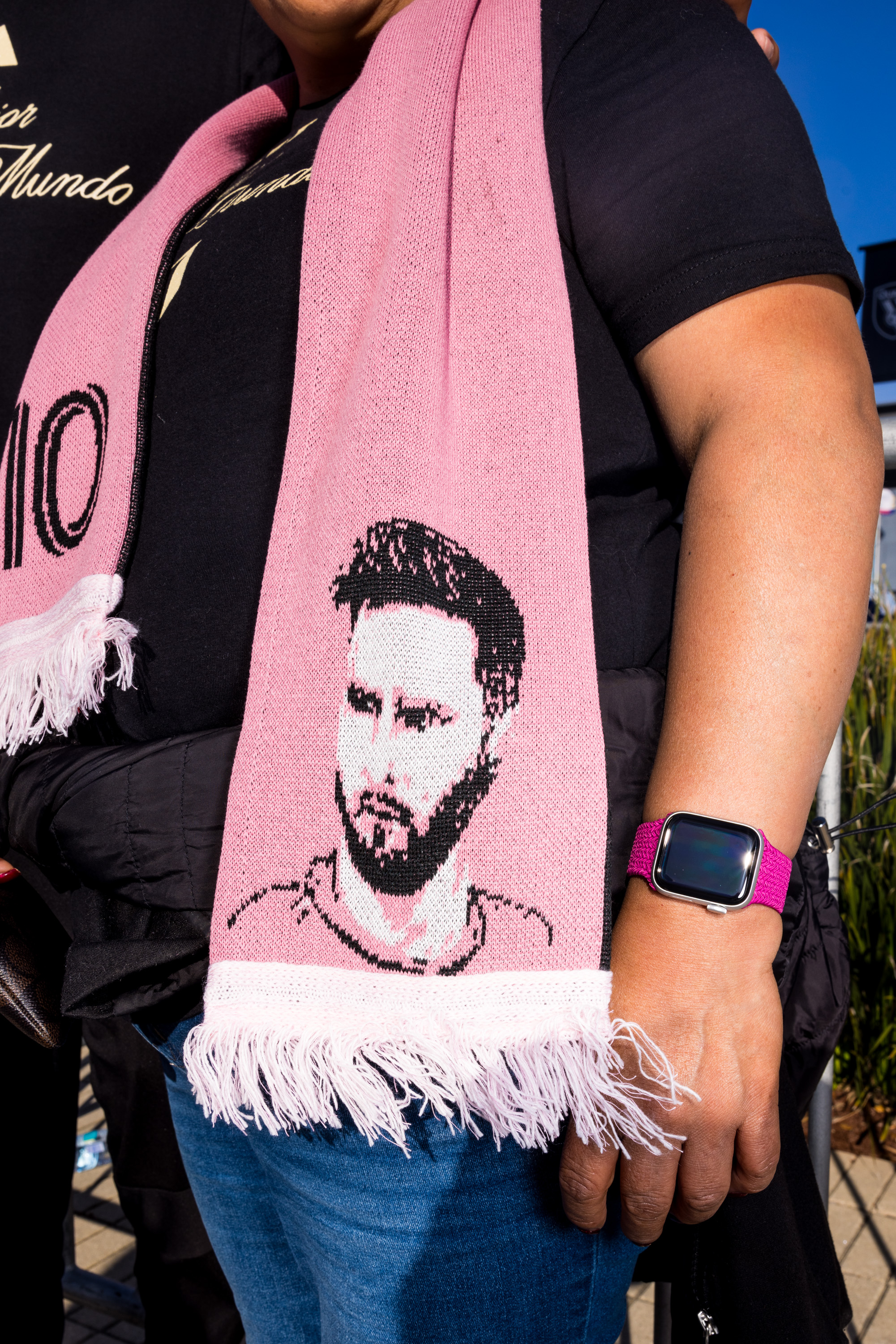 A person wearing a black shirt and a pink scarf with a portrait on it stands with an arm showing a pink watch, and their hand is close to their jeans.