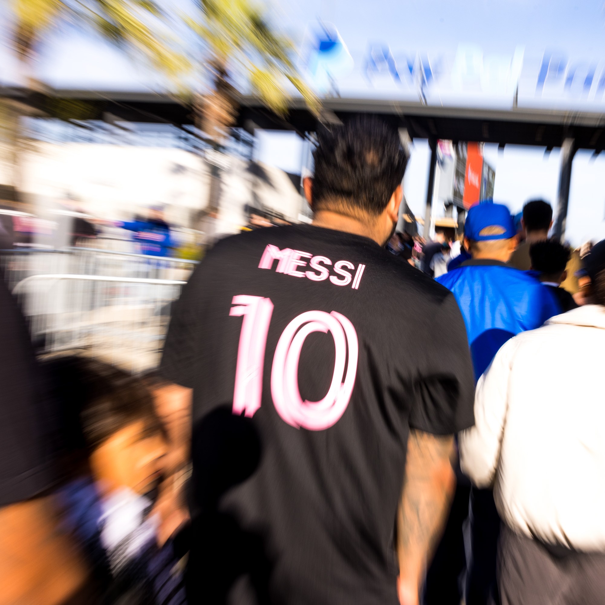 A crowd walks towards an entrance. One person in the foreground wears a black shirt with "MESSI 10" in pink letters. The image has a dynamic, blurred effect.