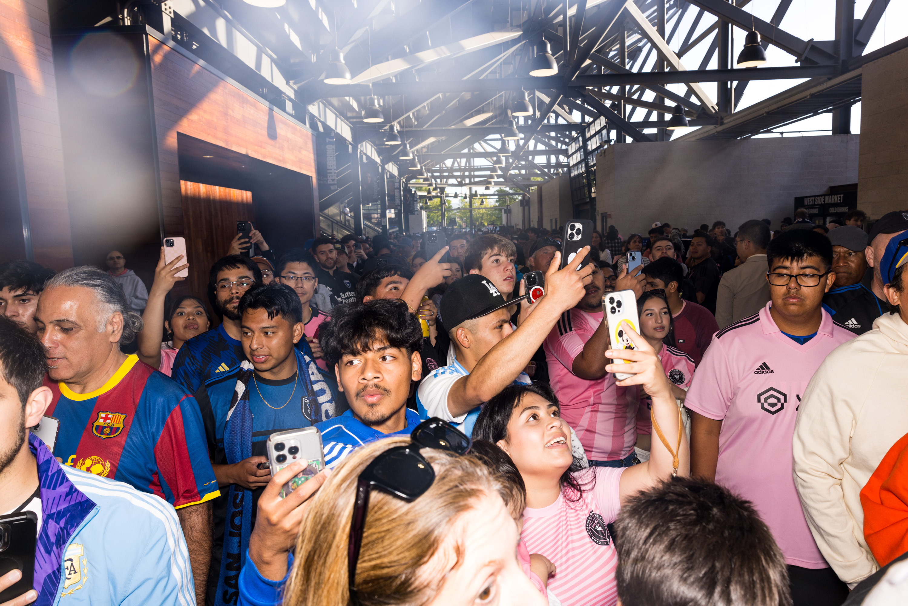 A bustling crowd with people holding up smartphones, wearing jerseys, and looking excited. The setting appears indoors with bright overhead lighting.