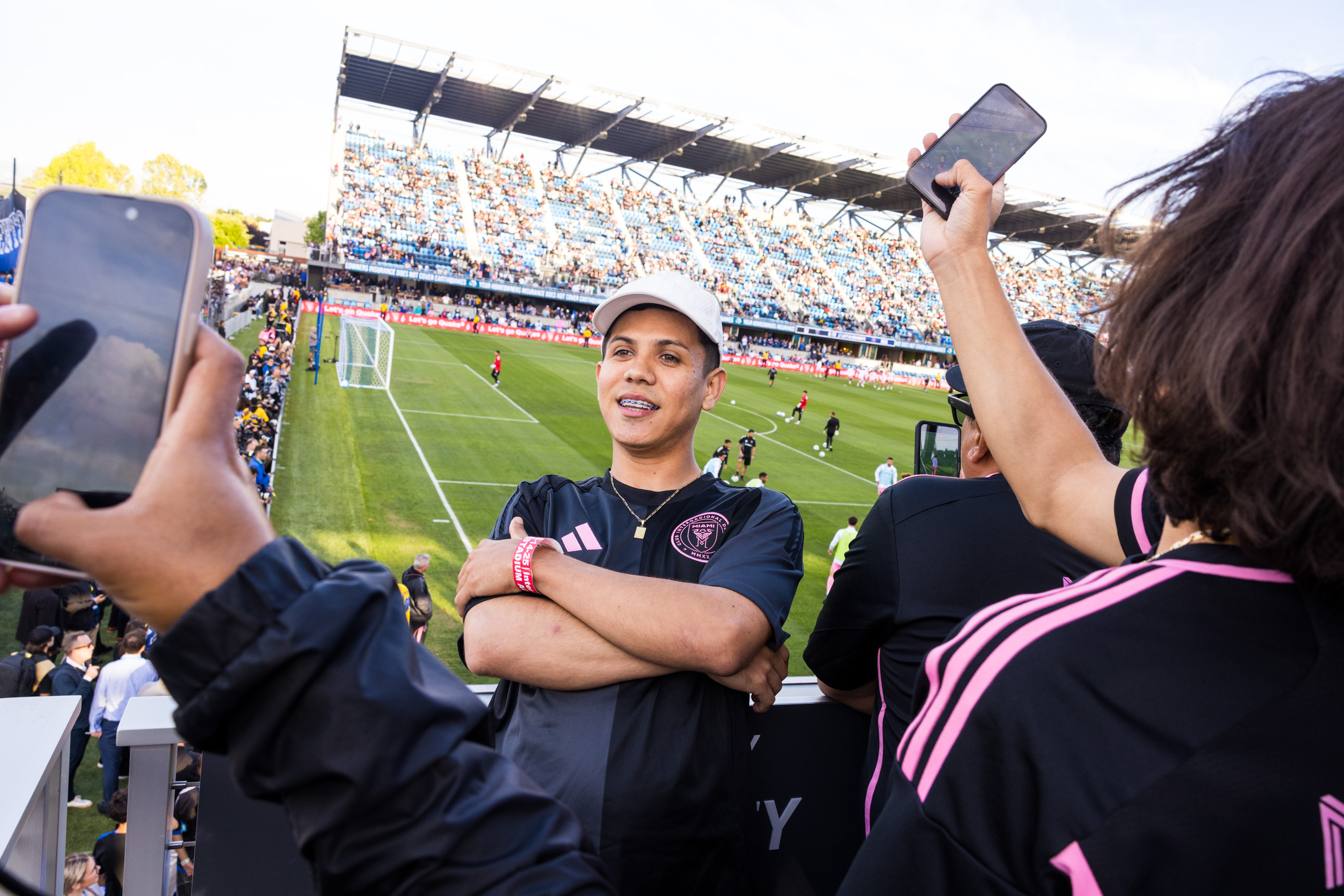 A man in a cap and black jersey stands smiling in a stadium, surrounded by people taking photos with their phones. A soccer match is happening in the background.