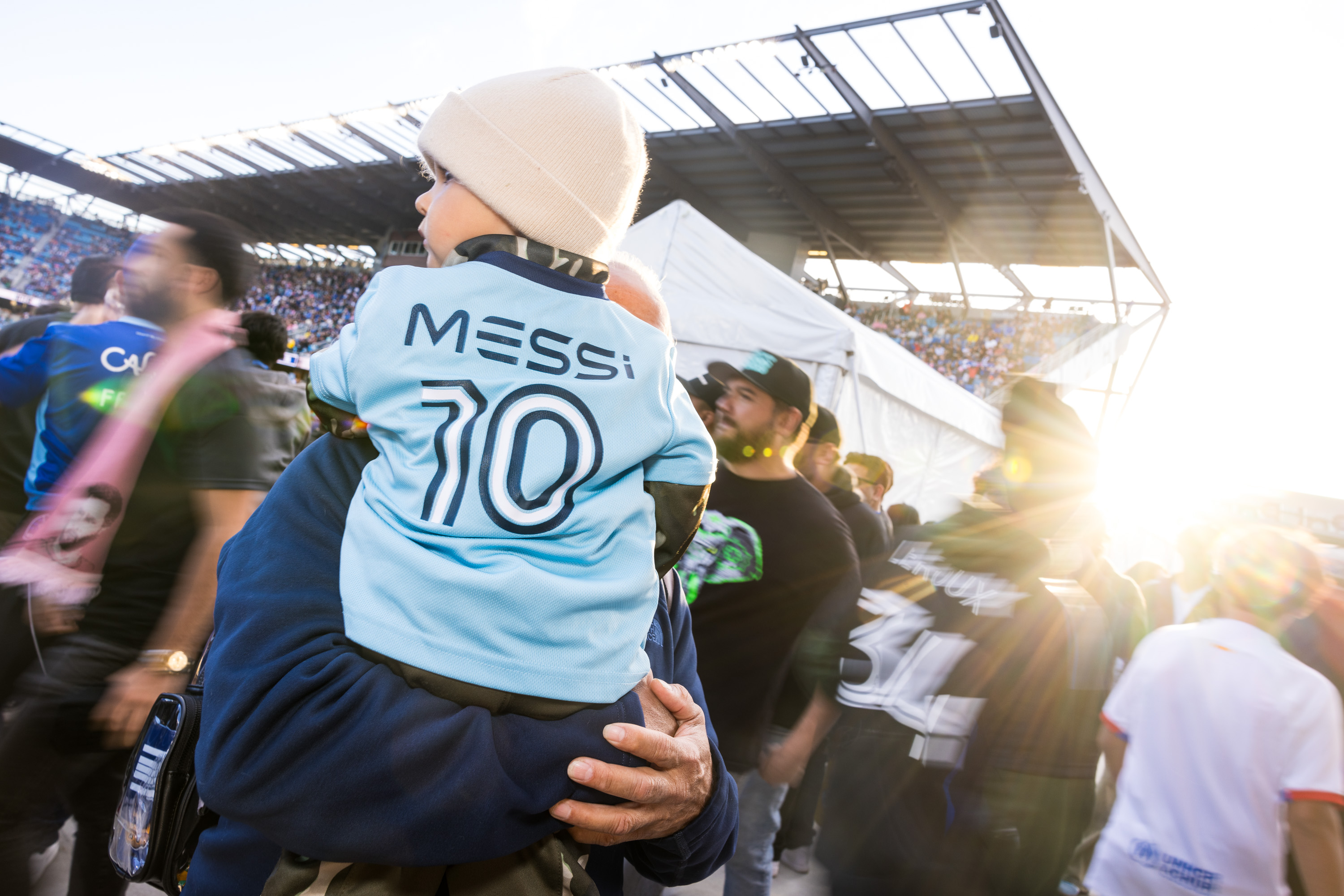 A young child in a &quot;Messi 10&quot; jersey is being held by an adult in a crowded stadium, with sunlight streaming in from the right side.