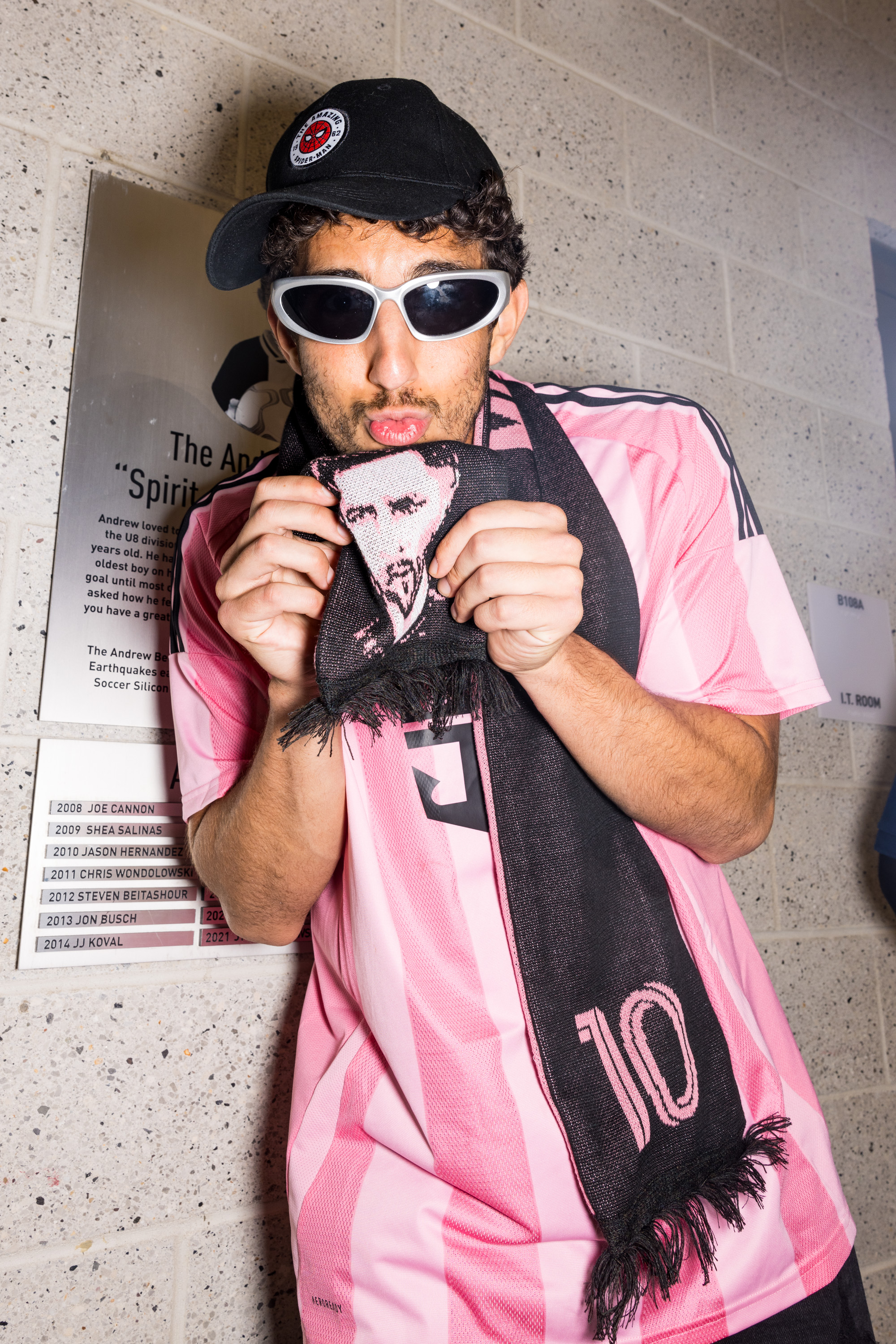 A person wearing a pink soccer jersey, black cap, white sunglasses, and holding a scarf with a face design, poses near a wall with commemorative plaques.