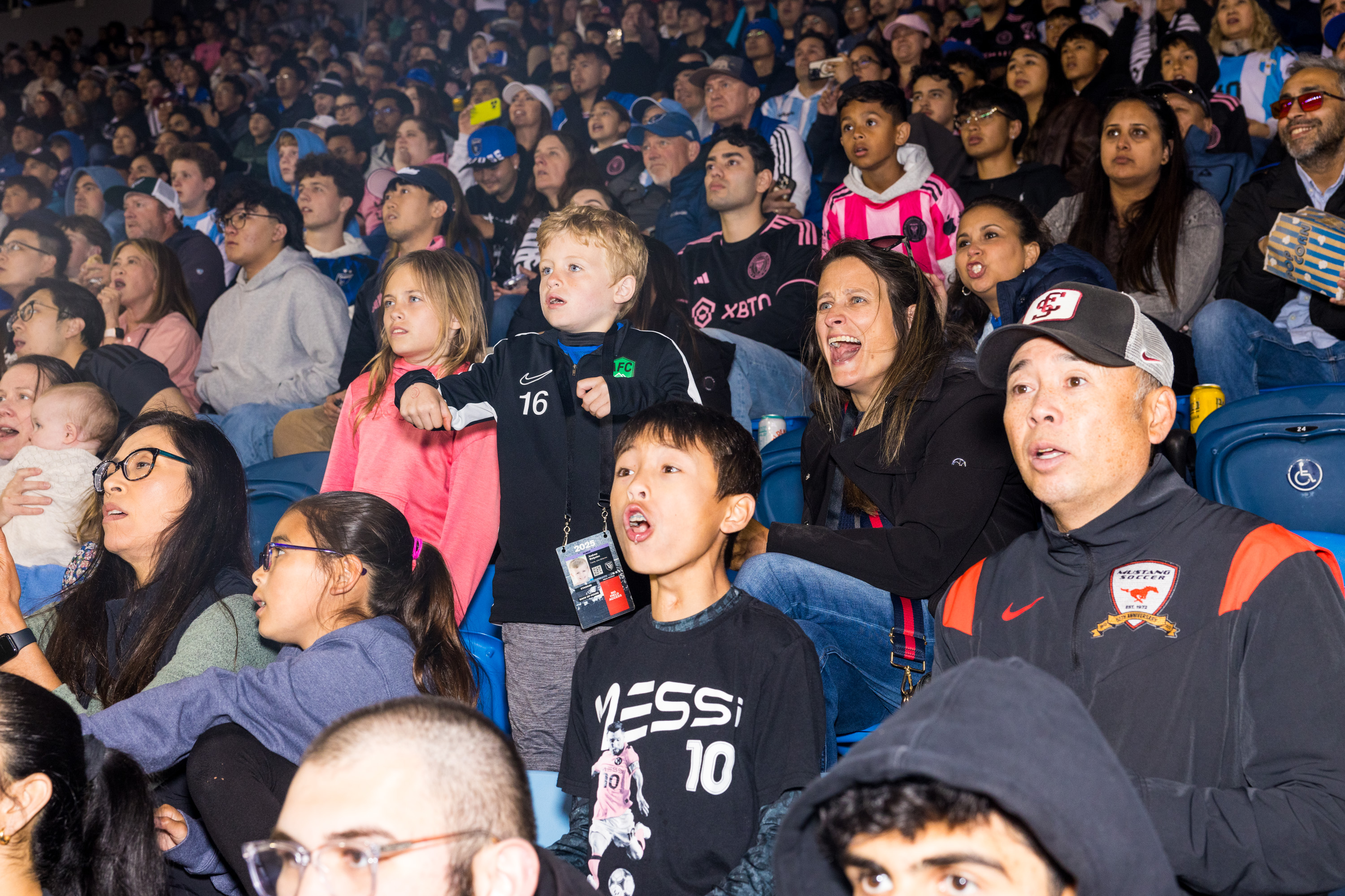 A diverse crowd of spectators, including children and adults, watch a game intently. They exhibit excitement and anticipation, with some wearing sports jerseys.