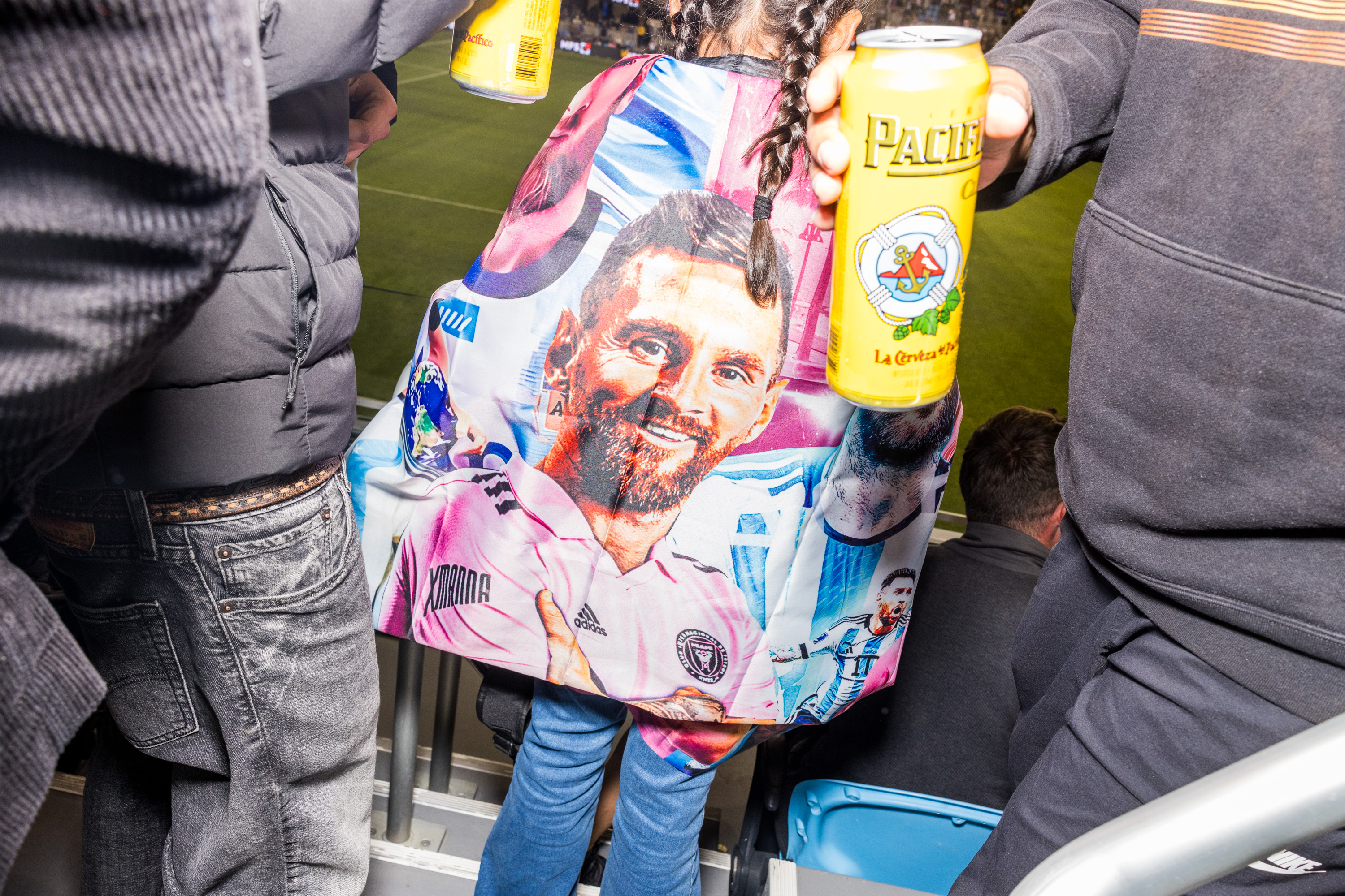 A person wears a colorful blanket featuring a soccer player's face, while others hold yellow beer cans at a sports event. The field is visible in the background.