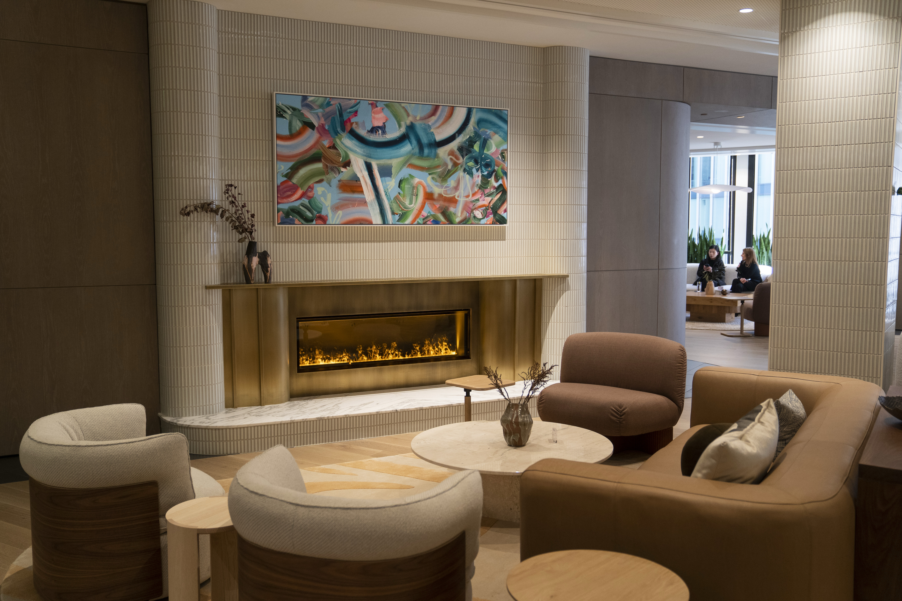 A cozy lounge features a modern fireplace, abstract art above, plush seating, and a round table with a decorative vase. Two people are seated in the background.