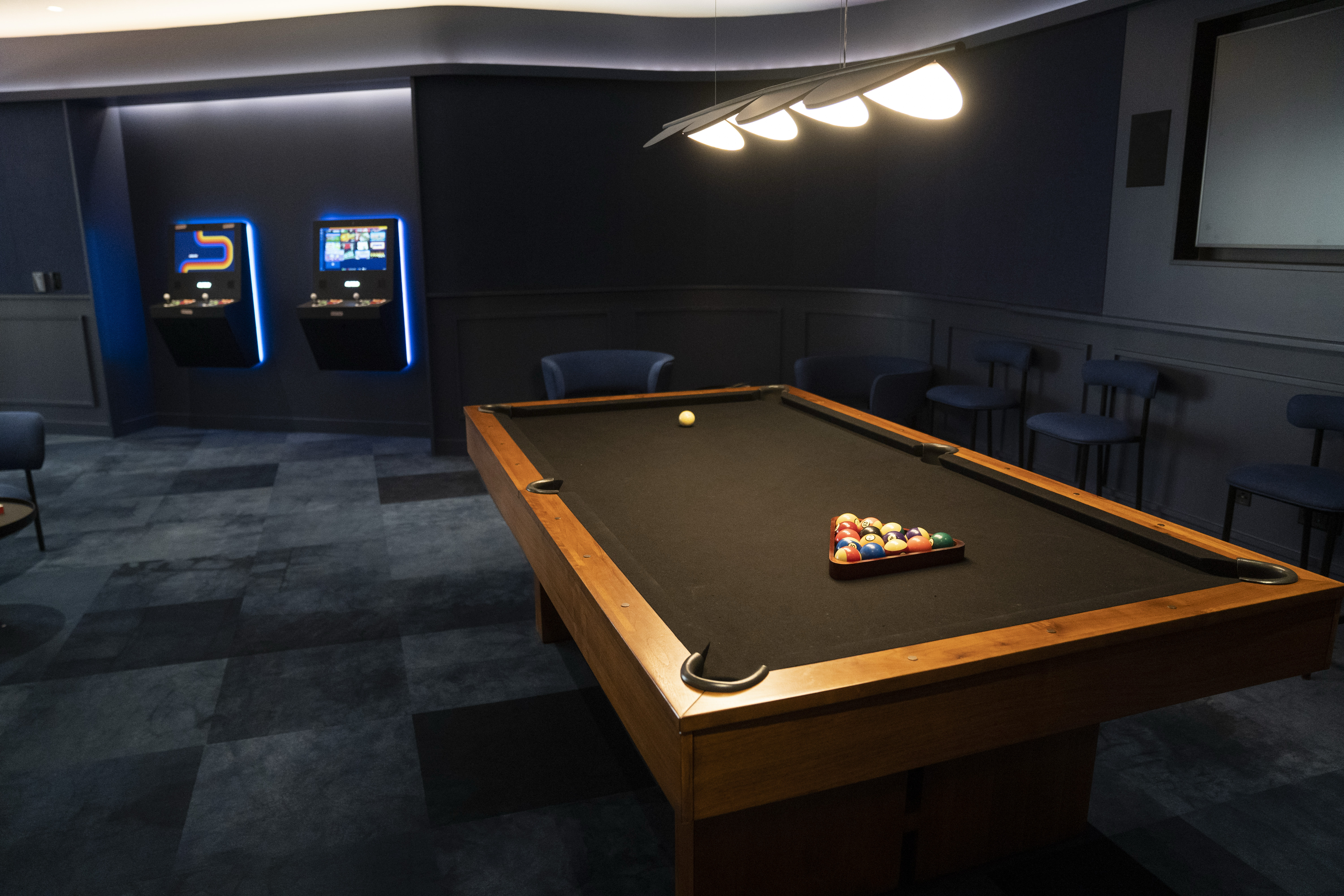 A modern game room features a pool table with balls arranged. Two arcade machines glow in the corner, and several bar stools are placed against dark-paneled walls.
