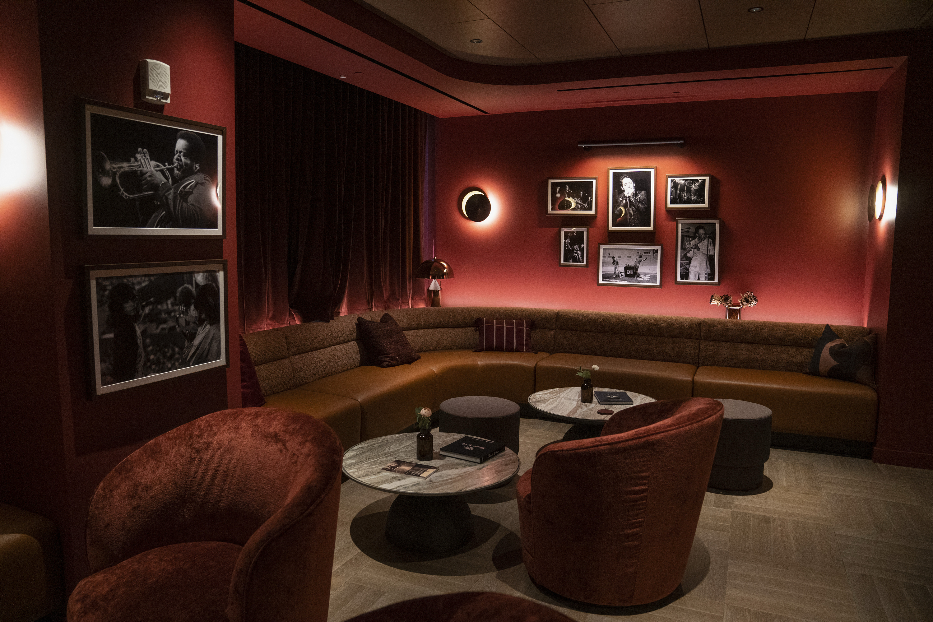 The image shows a cozy lounge with red hues, velvet chairs, and a curved sofa. Black-and-white photos adorn the walls, and soft lighting creates a warm ambiance.