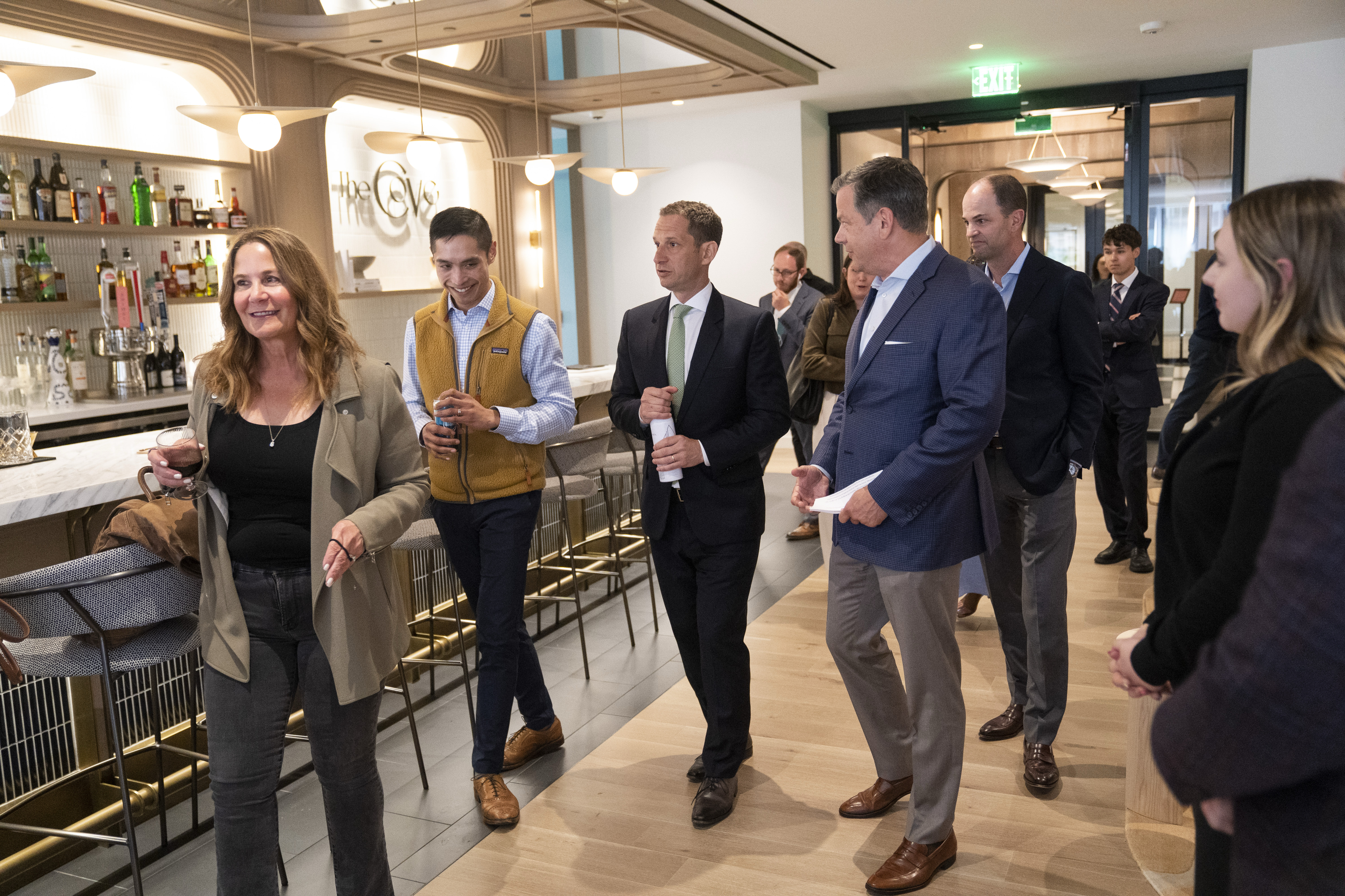 A group of people walks through a stylish bar area, dressed in business attire. They appear engaged in conversation, with drinks in hand.