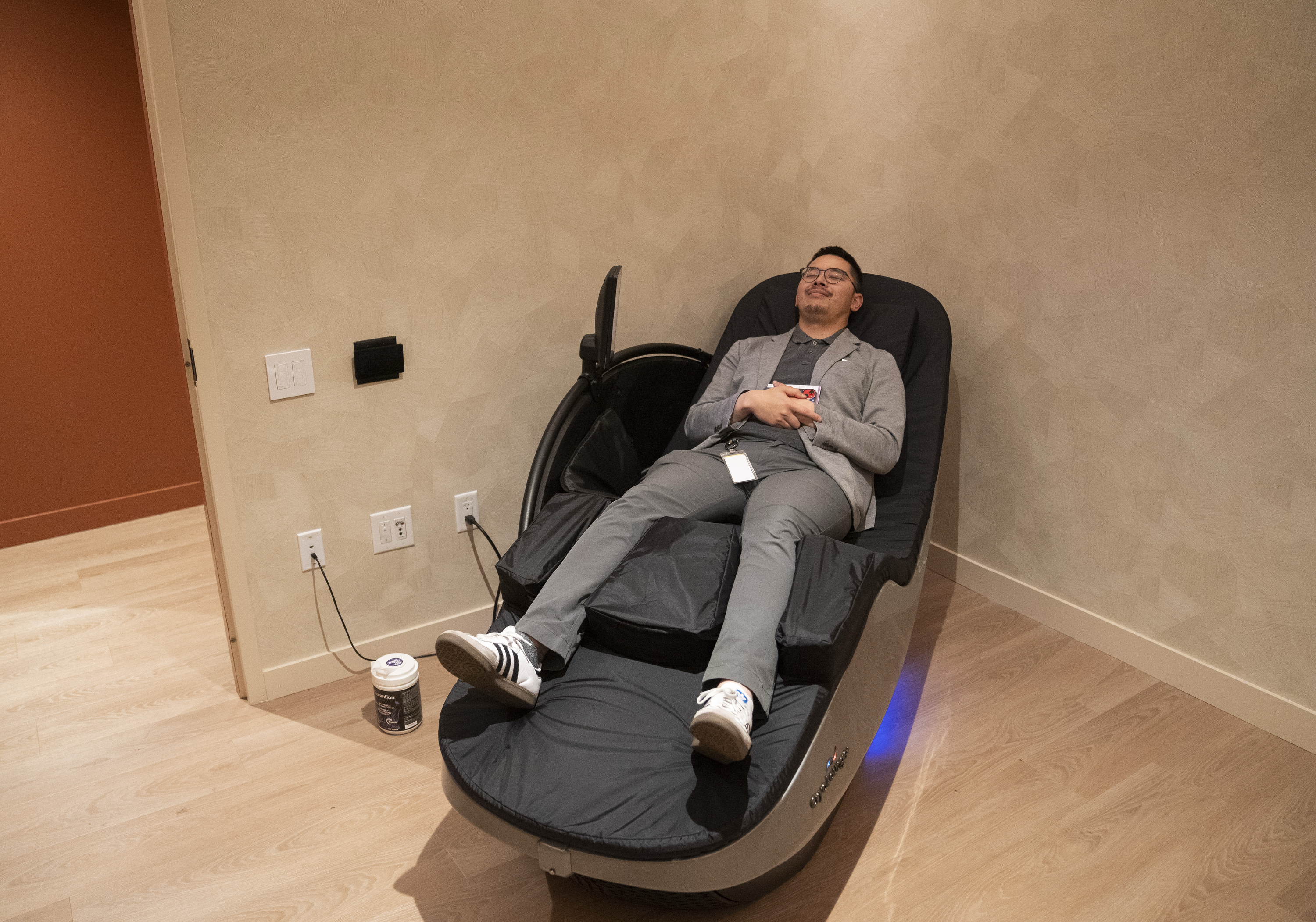 A person is relaxing in a reclining massage chair wearing casual clothes. The room has wood flooring and a small container on the floor next to the chair.