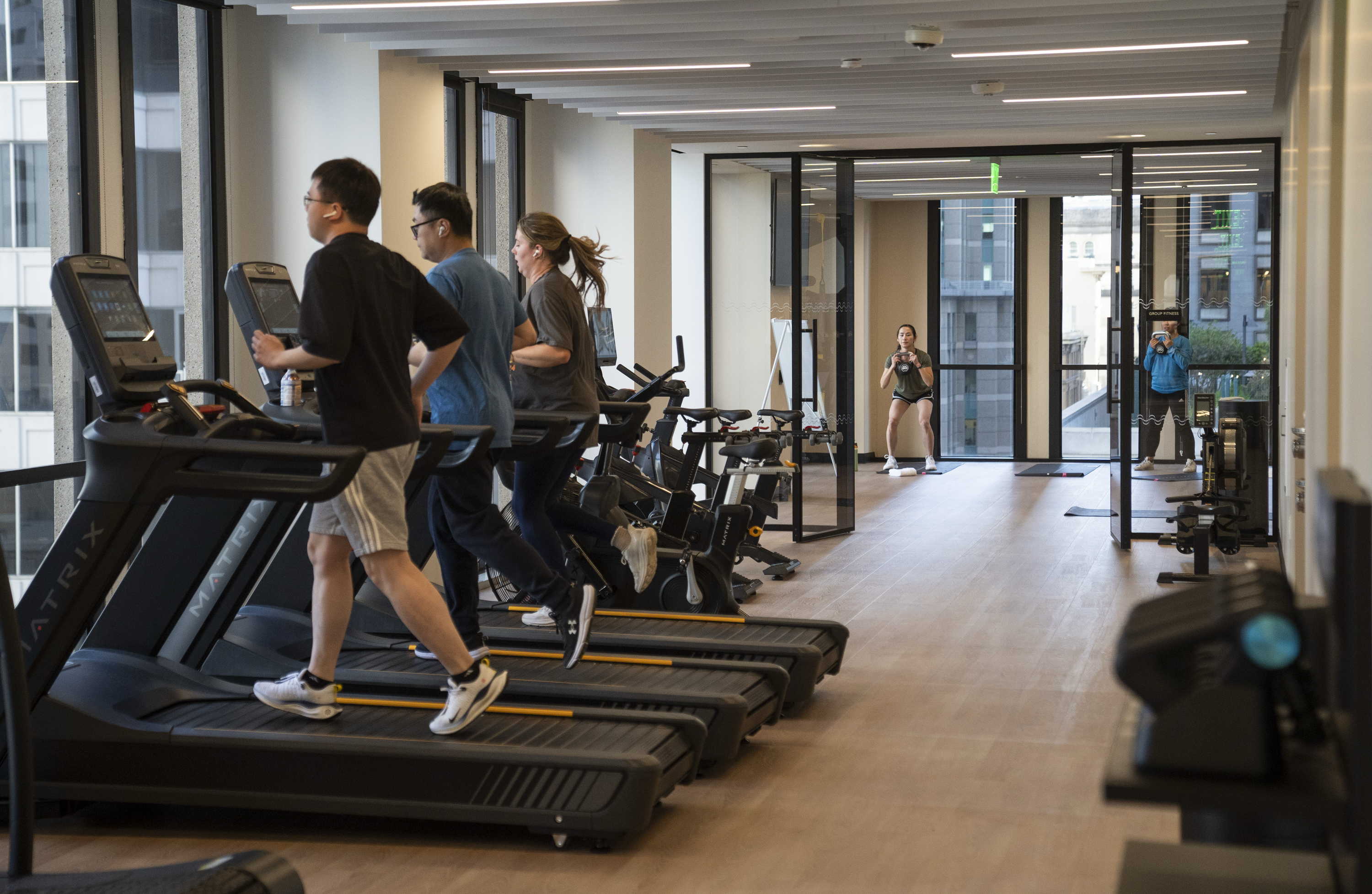 Several people are running on treadmills in a gym, with others exercising in the background. The gym has large windows and modern equipment.