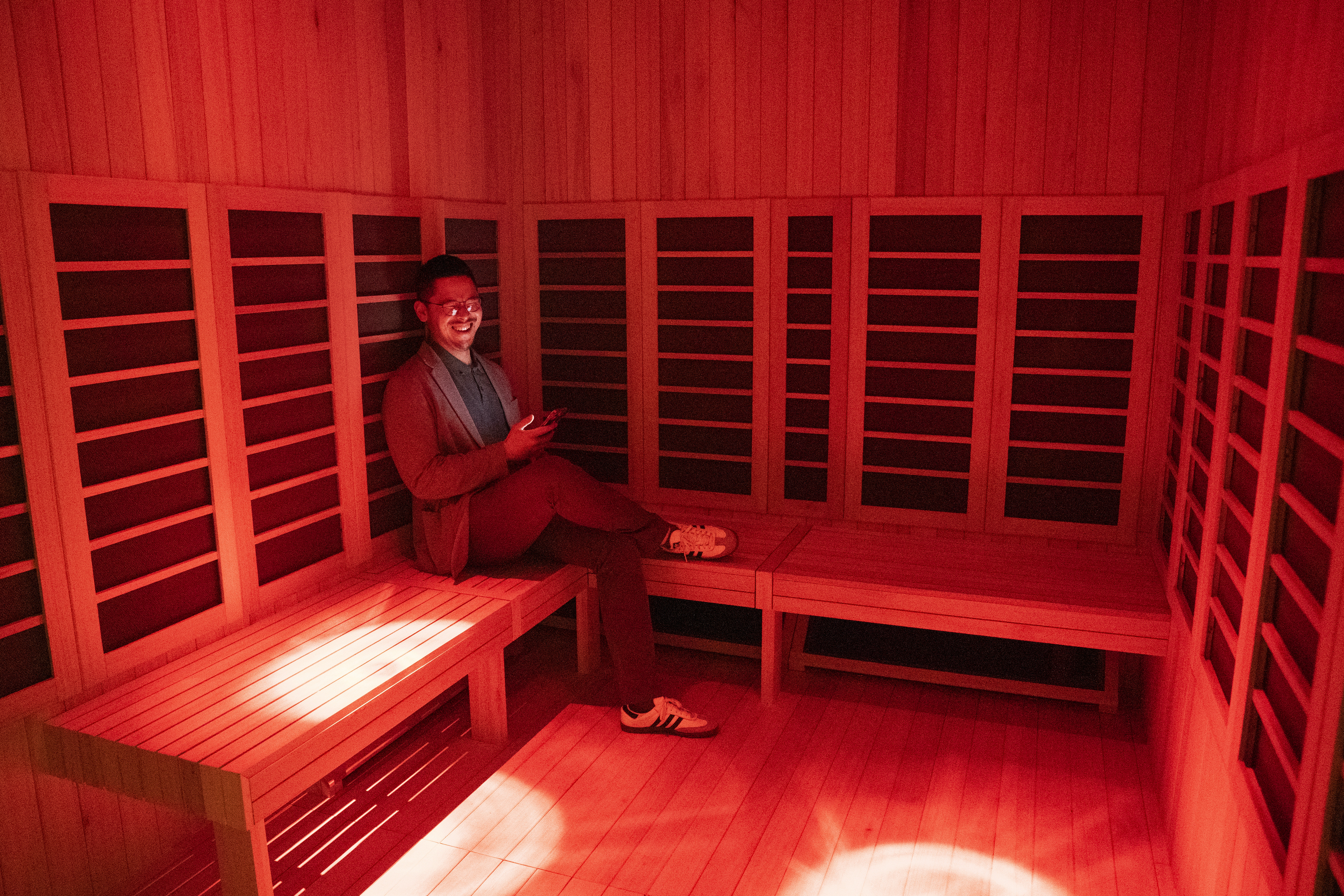 A person sits smiling in a red-lit wooden sauna, holding a phone. They wear glasses, a sweater, and sneakers, and the room has benches with a warm ambiance.