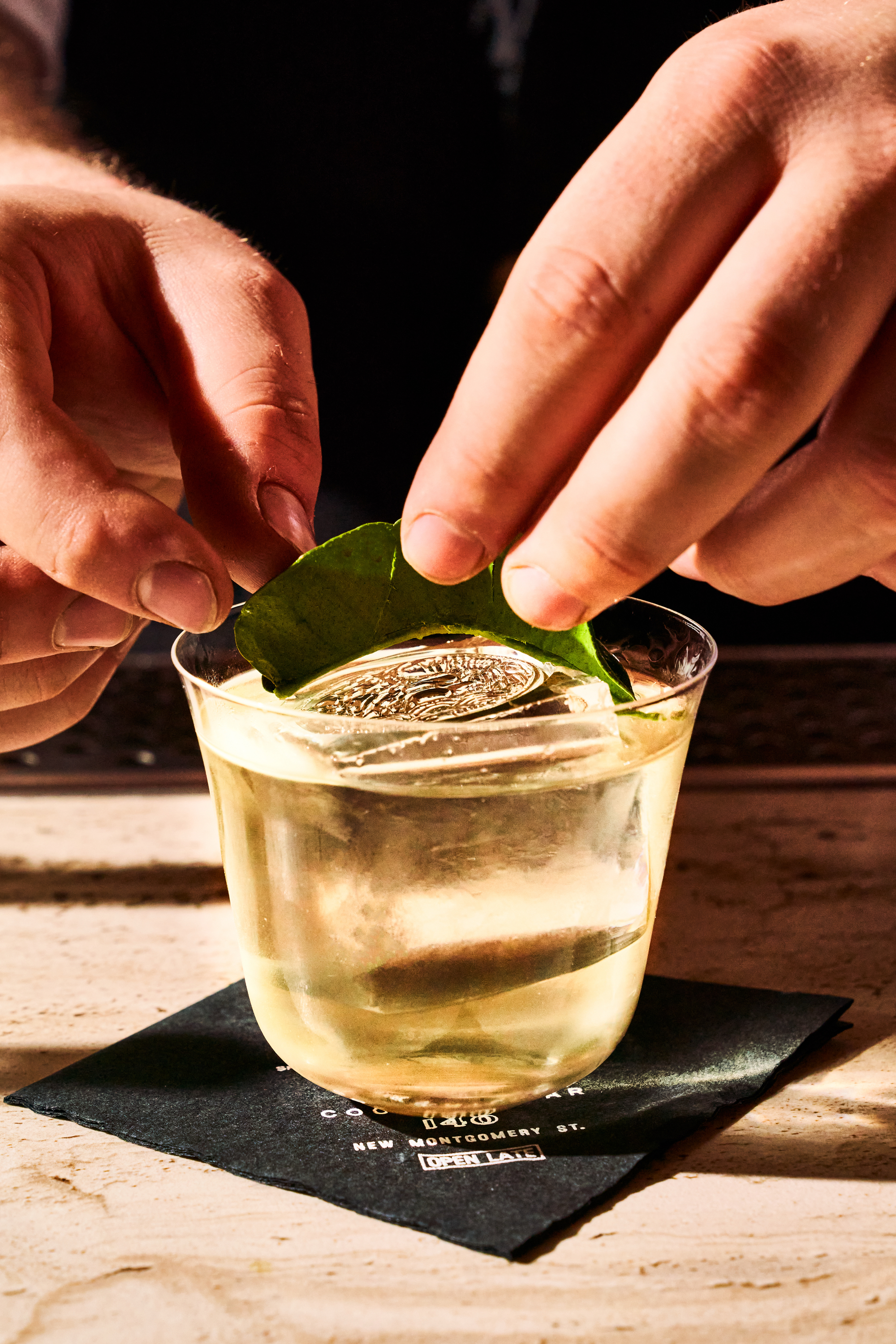 A person is placing a green leaf on a clear cocktail in a glass with a coin-like garnish, set on a dark napkin on a light surface.