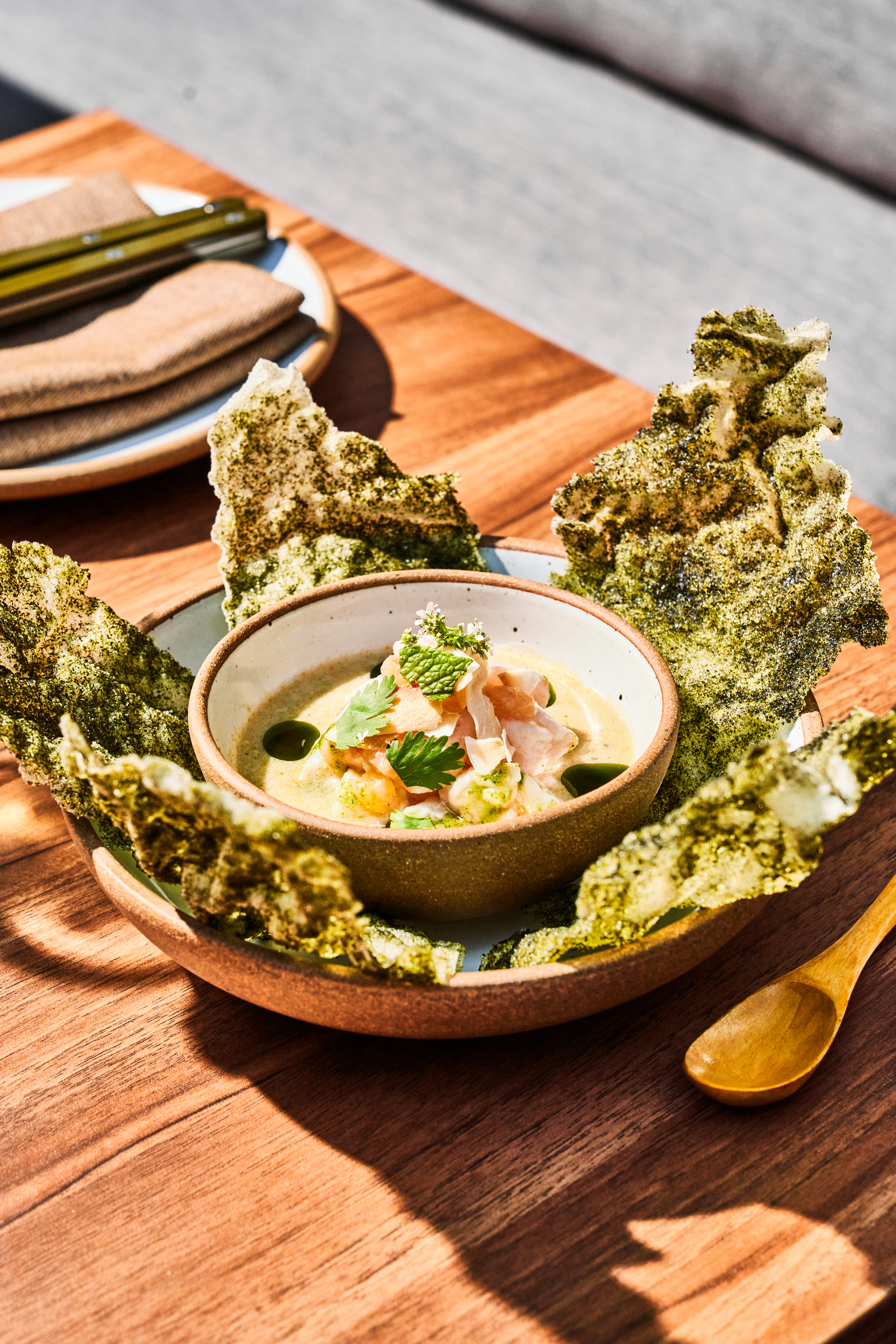 A ceramic bowl holds a creamy dish topped with herbs, surrounded by large, crispy seaweed on a wooden table, next to a wooden spoon and folded napkins on a plate.