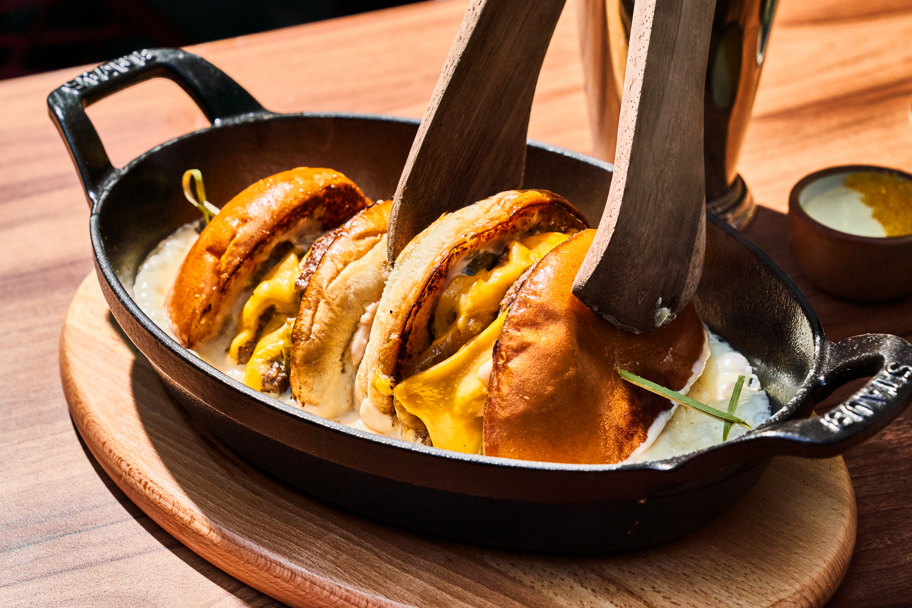 Three cheeseburgers are stacked in a black skillet with melted cheese, secured by wooden boards, on a wooden surface with a small bowl nearby.